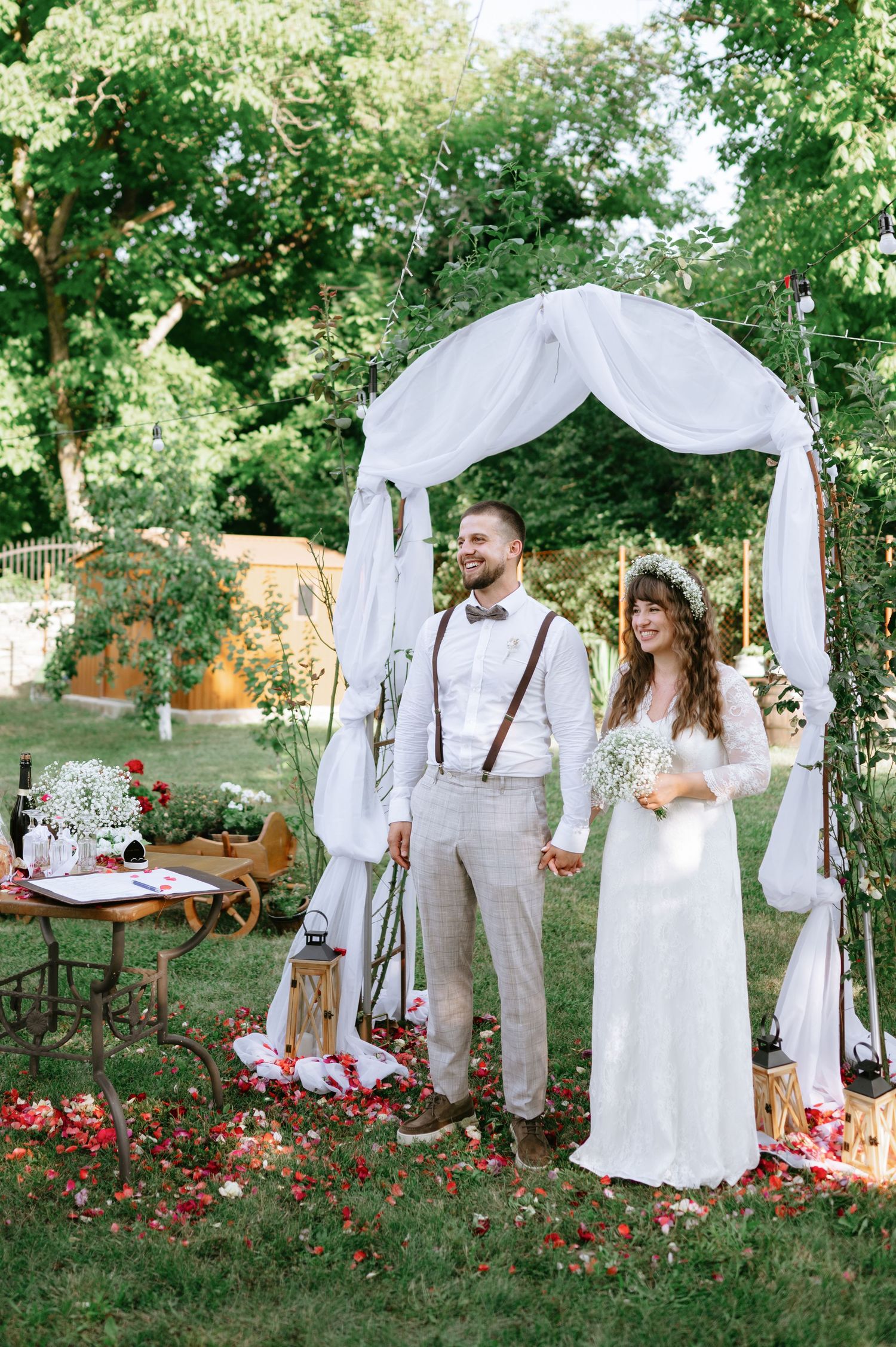 Rustic outdoor wedding scene with white fabric arch and vintage decor in garden setting.
