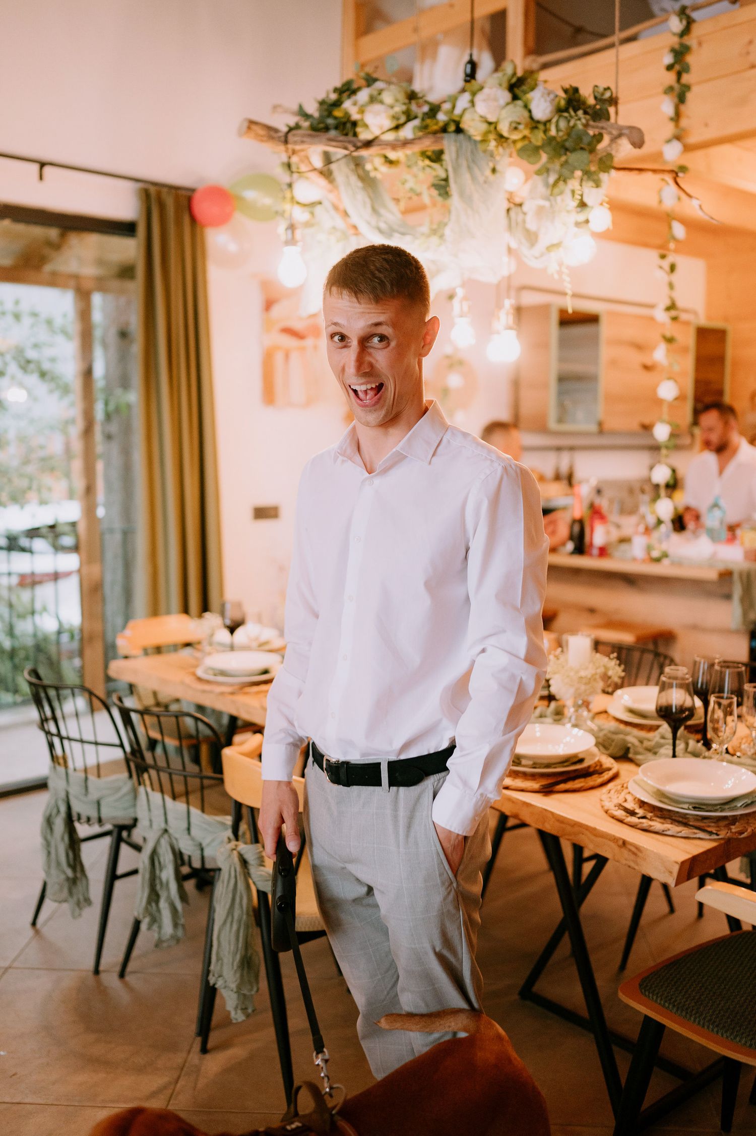 A person in formal attire smiles enthusiastically in front of an elegantly decorated dining table.