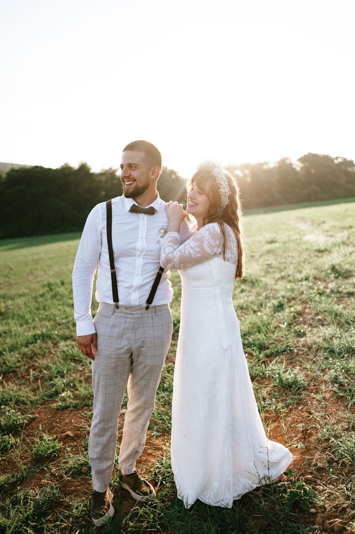 Romantic wedding couple embraces in sunny field wearing white dress and suspenders.