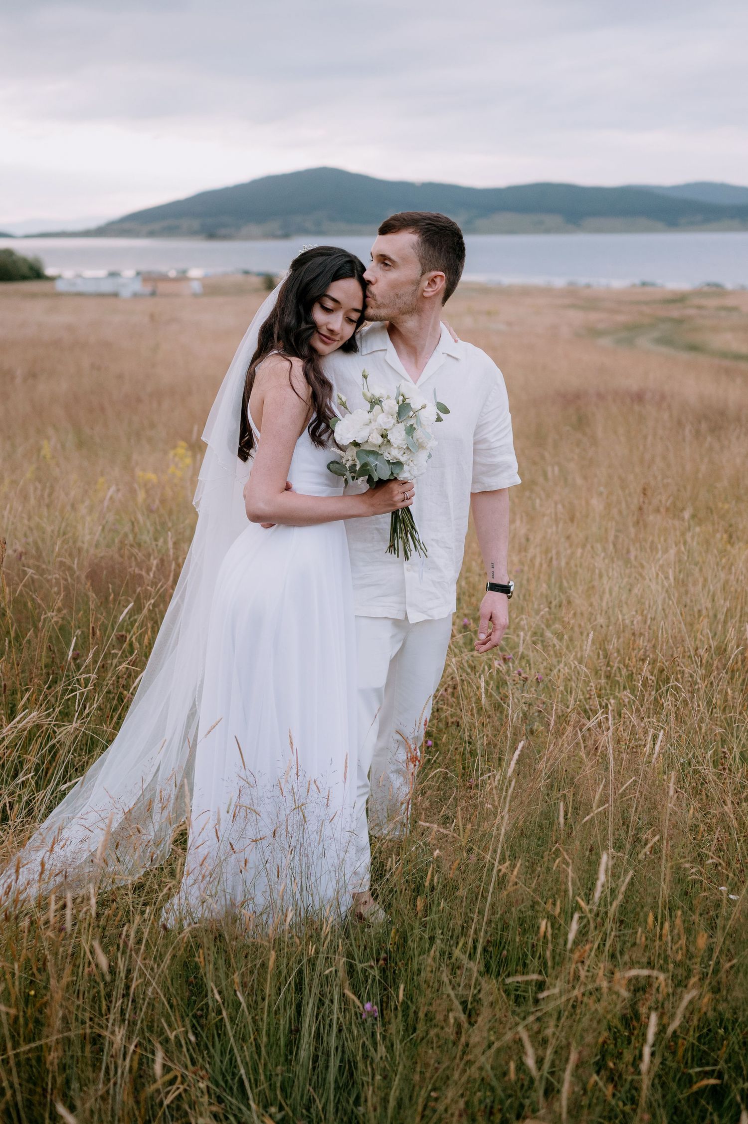 A romantic wedding couple embraces in a golden field with mountains and water in the distance.