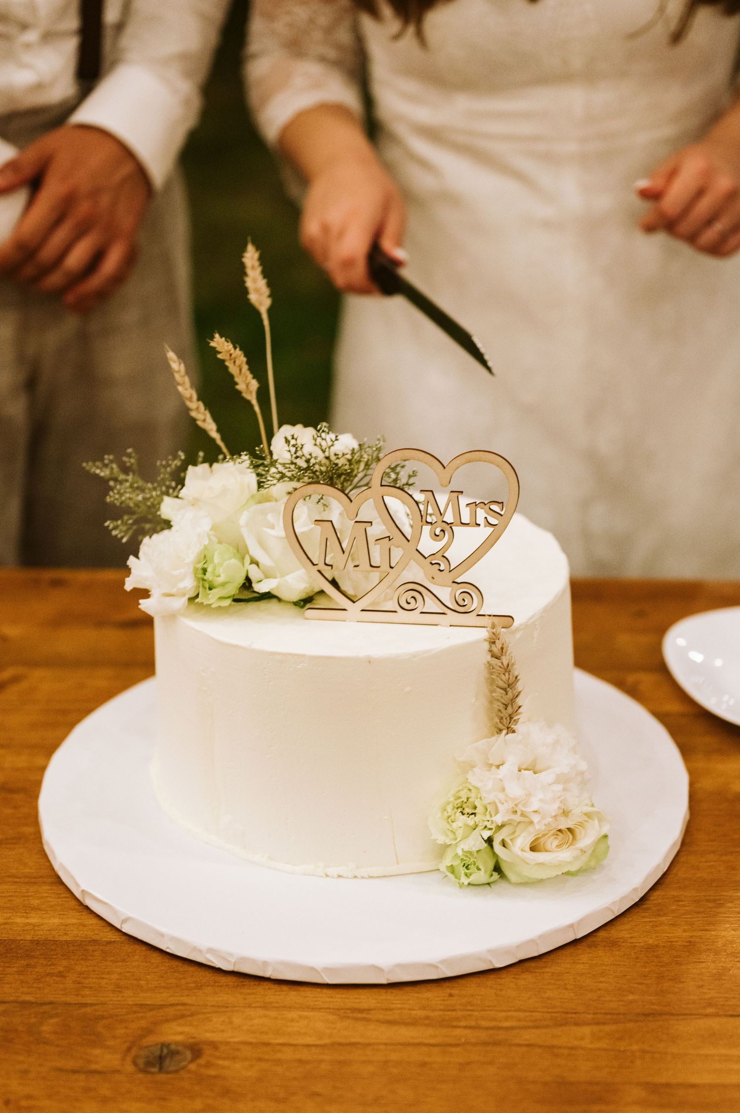 White wedding cake decorated with floral accents and a Mr & Mrs wooden topper on rustic table.