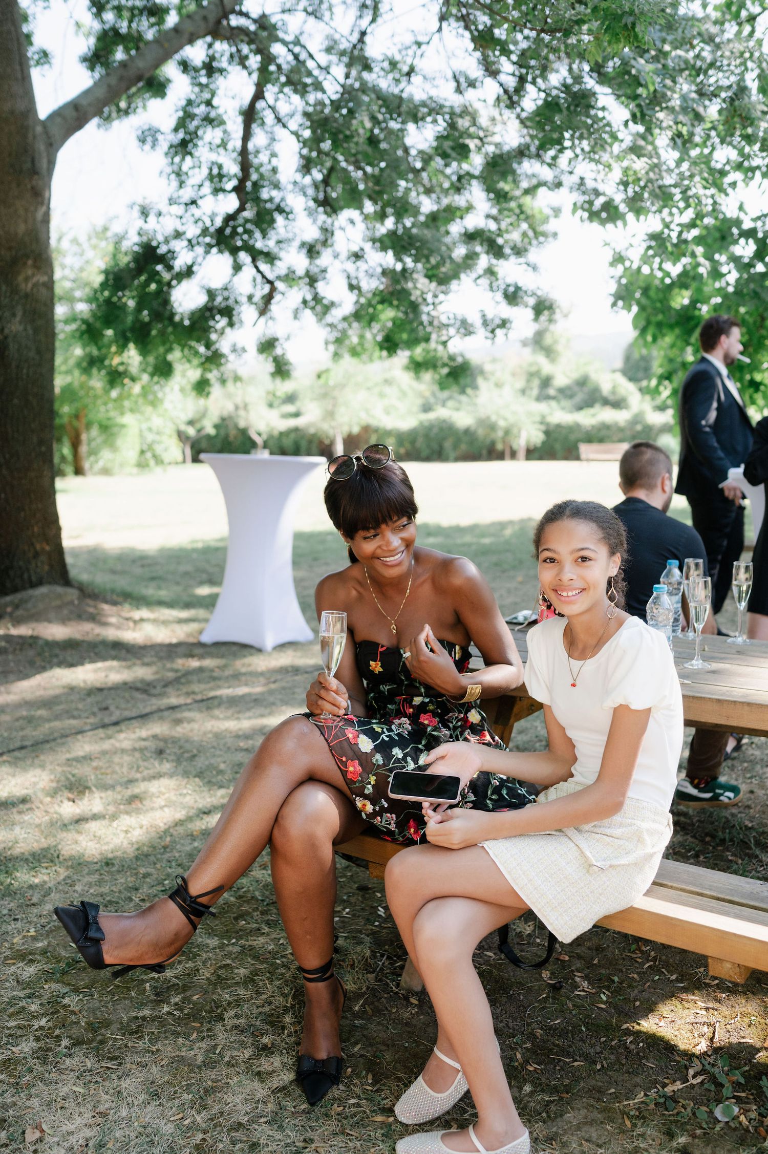 Two friends share a laugh while sitting on a wooden bench in a shaded outdoor setting.