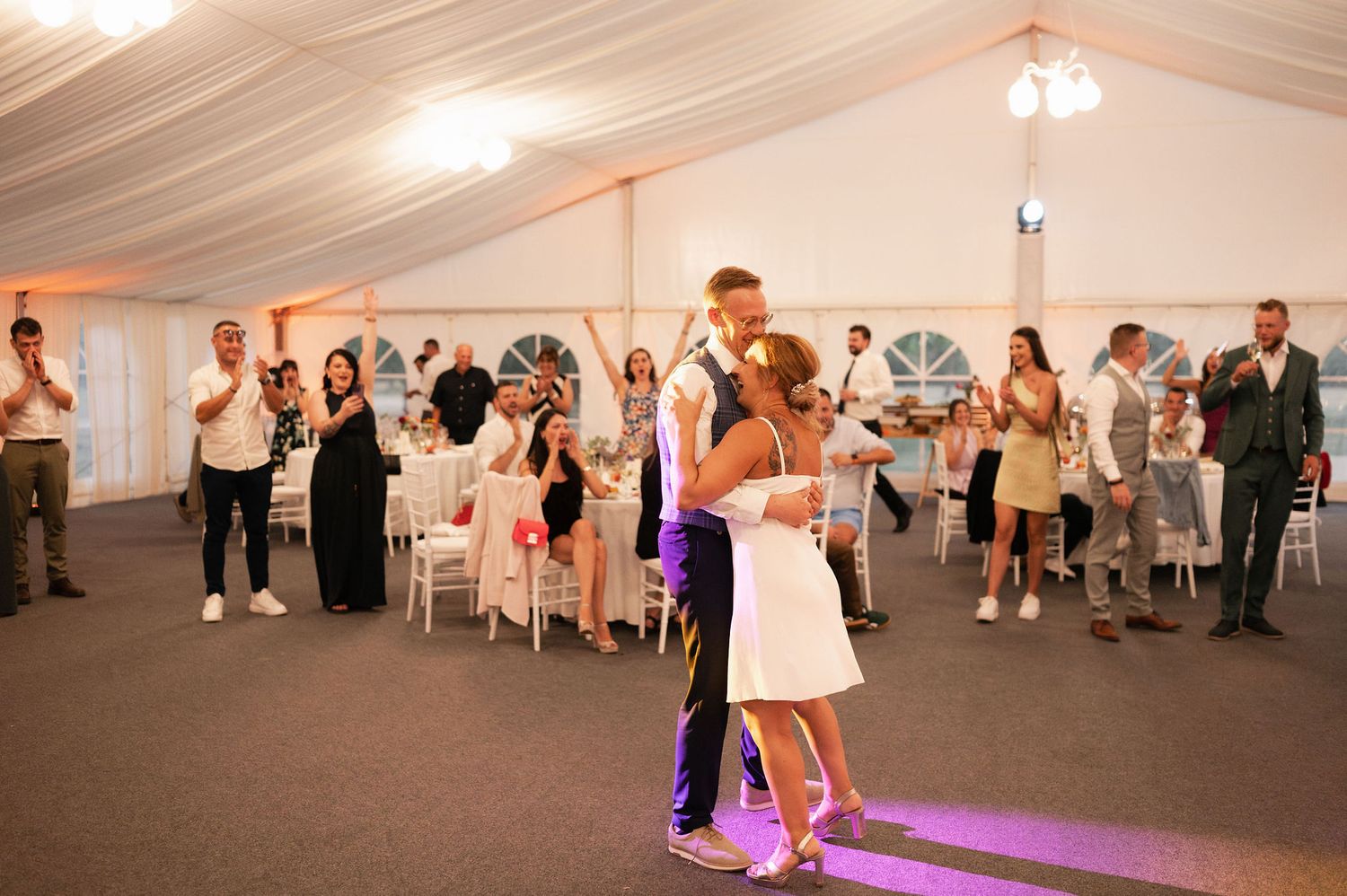 Wedding guests dance and celebrate on a purple-lit dance floor inside a white tent reception venue.