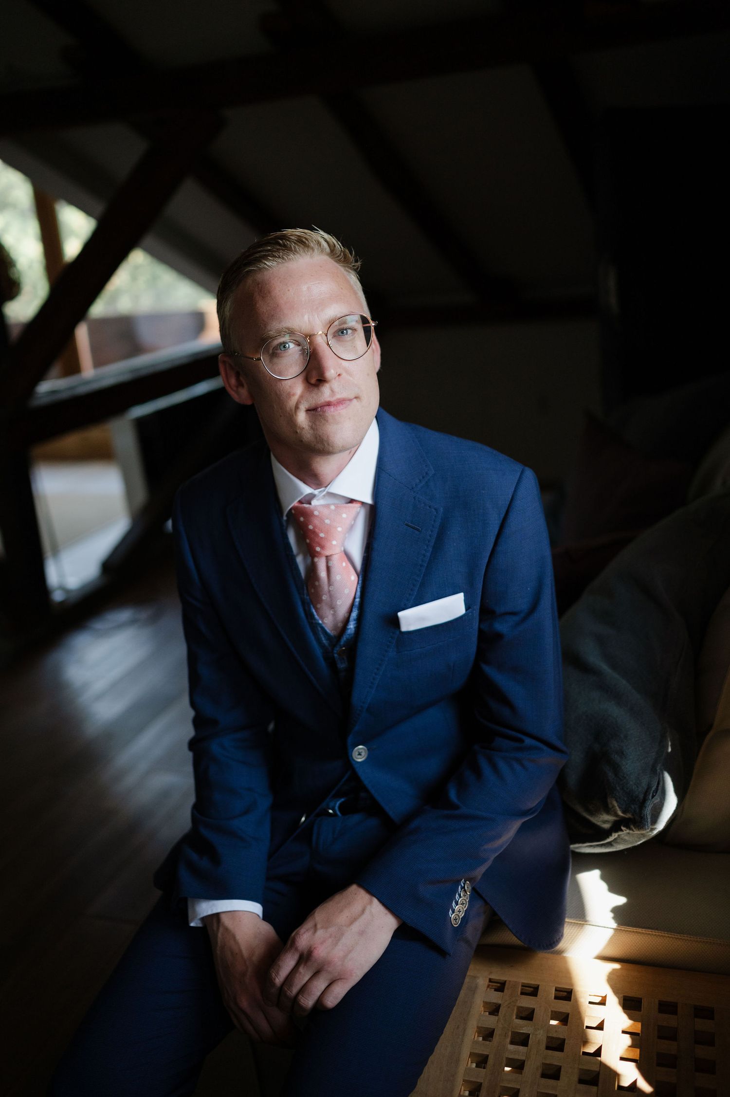 Dramatic lighting captures someone in a navy suit sitting in a dark room.