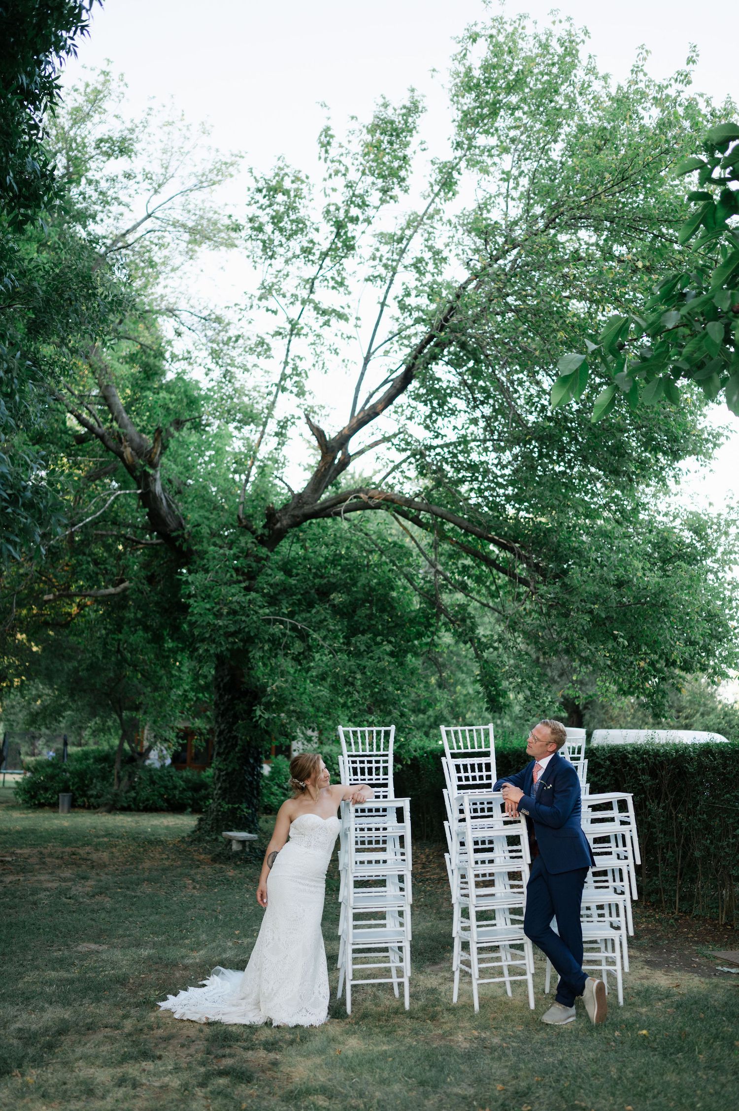 A wedding couple moves between white chairs set up in a garden venue.