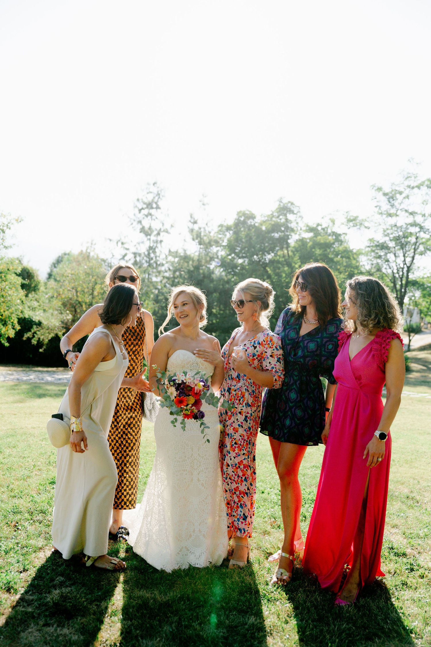 Bridesmaids in an array of colorful dresses stand together in bright sunlight.