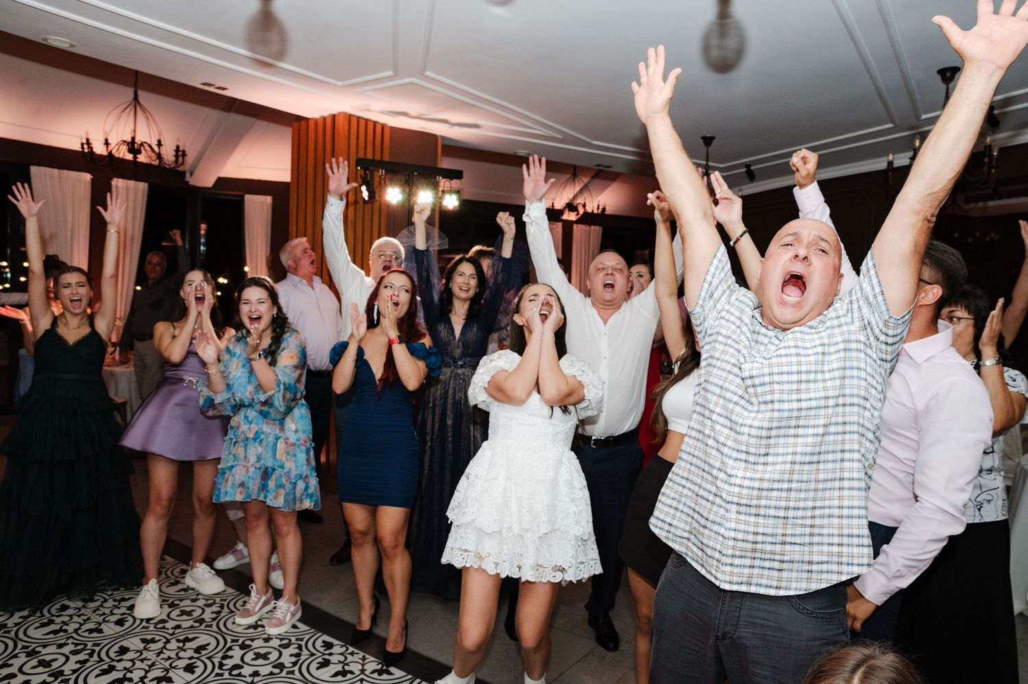 Crowd of wedding guests with raised hands dancing enthusiastically under indoor lighting.