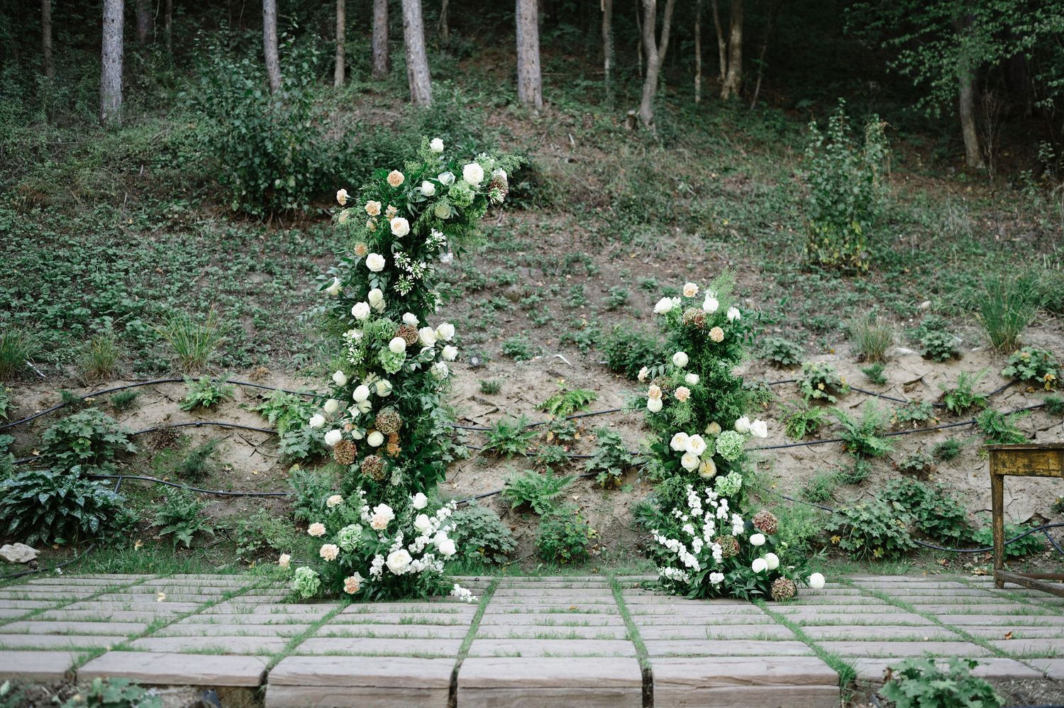 Garden wedding ceremony space with white floral arrangements and natural woodland backdrop.