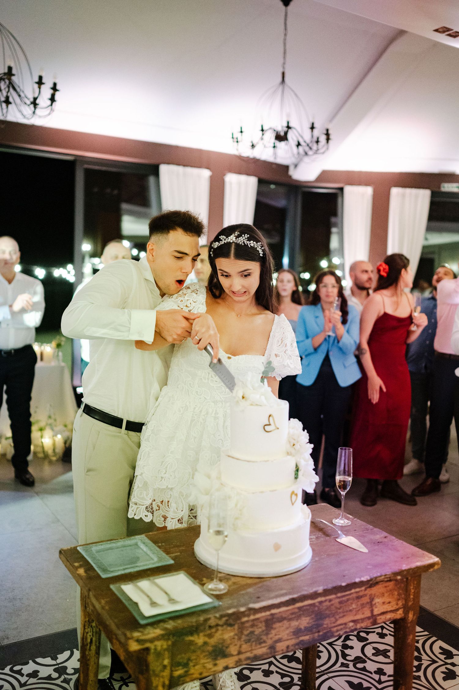 Newlyweds share intimate cake cutting moment while guests watch in background.