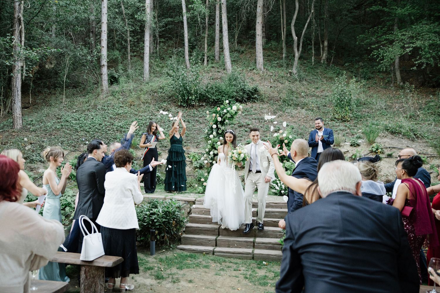 Wedding guests seated on wooden benches in a forest amphitheater setting.