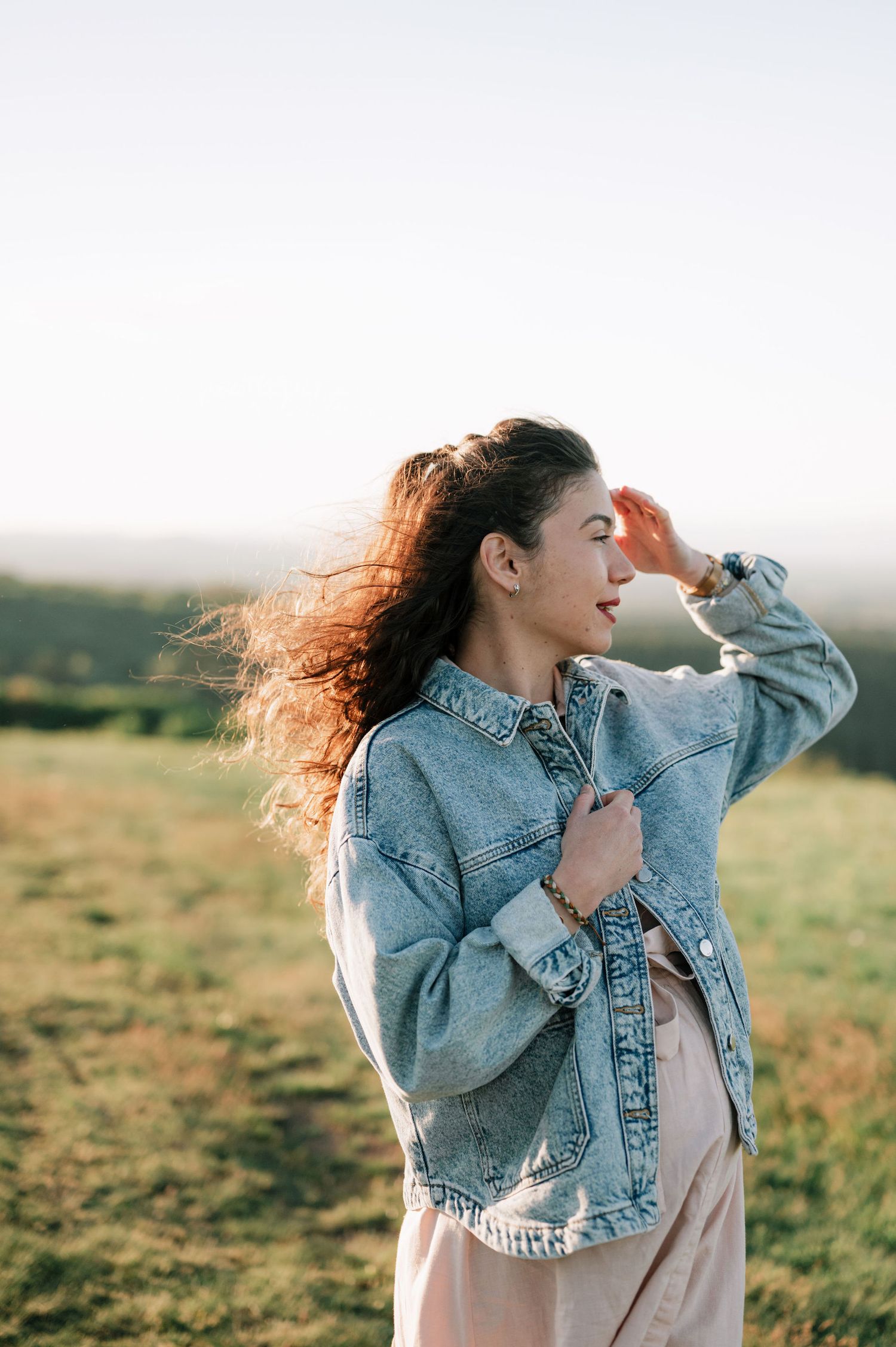 A person in a denim jacket stands in a grassy field at sunset with their hair flowing in the wind.