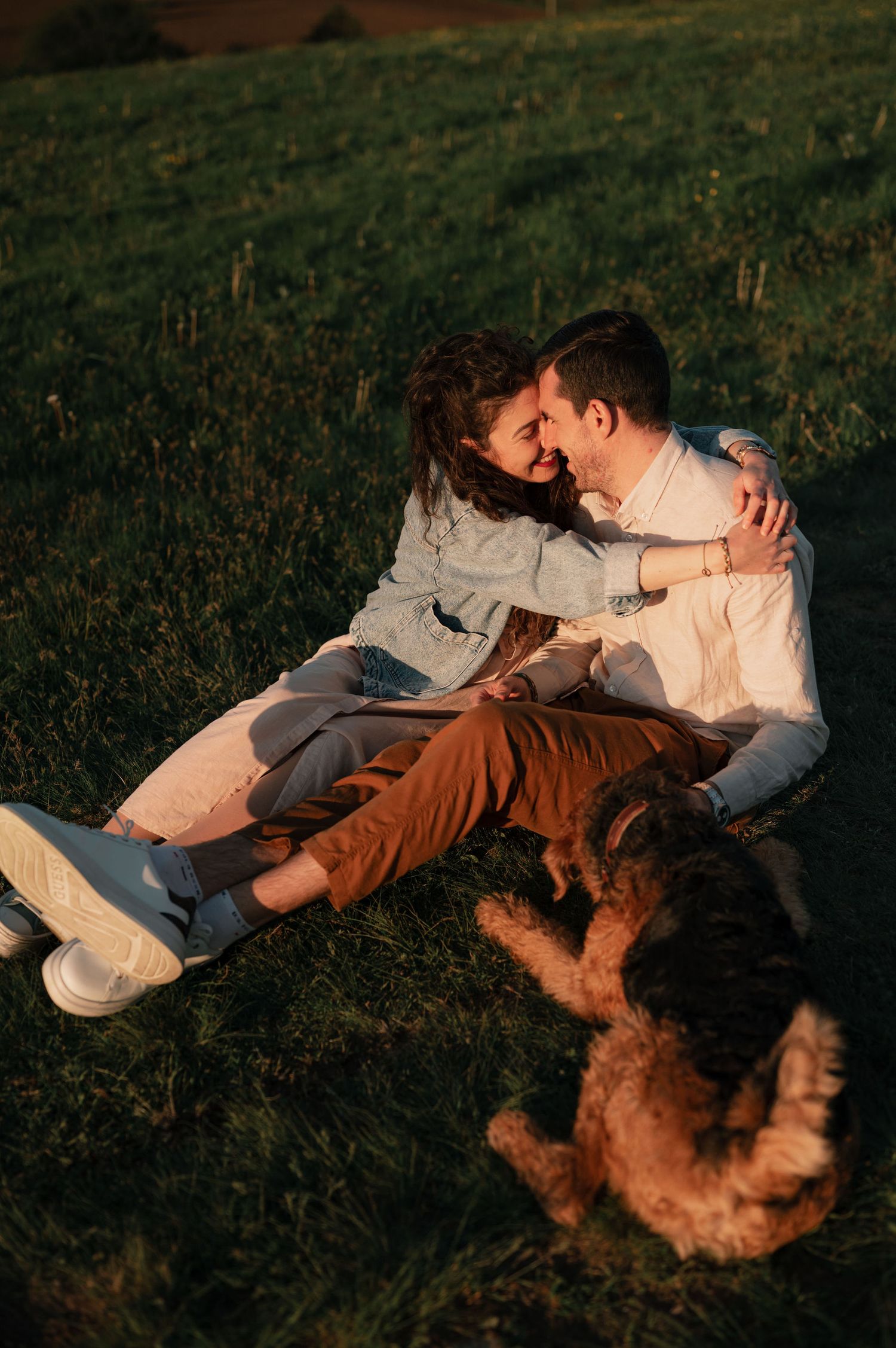 Couple lounges on hillside with their dog during golden hour sunset.