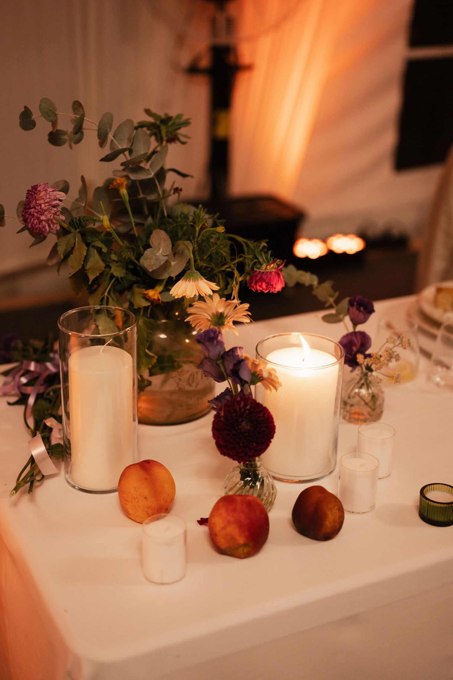 Elegant table setting with lit white candles and small floral arrangements in fruit centerpieces creating warm ambiance.