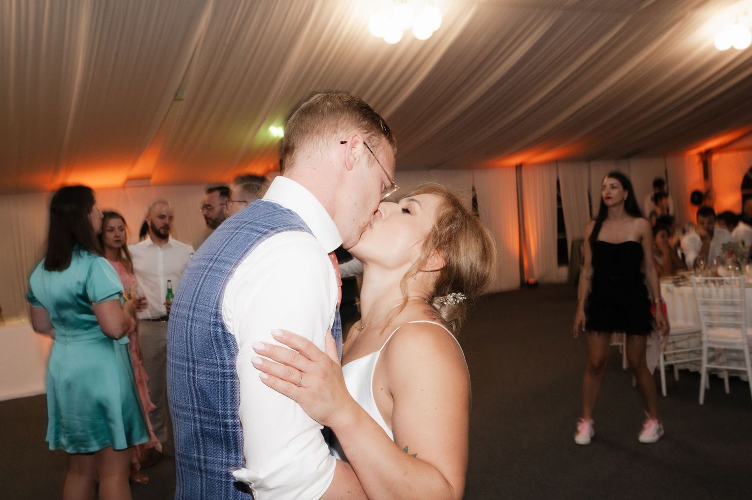 A couple shares a romantic kiss on the dance floor at a wedding reception with guests and orange lighting in the background.
