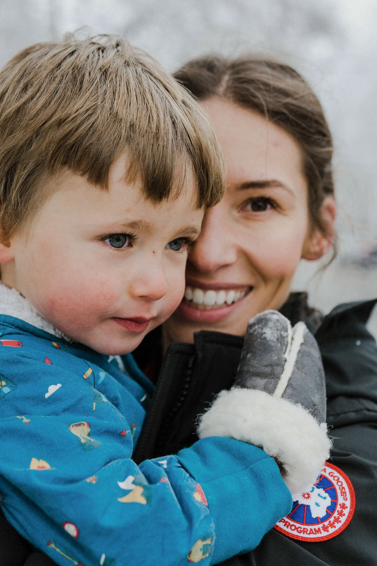 A child in a blue patterned coat and winter gloves shares a close moment with an adult wearing a Canada Goose jacket.