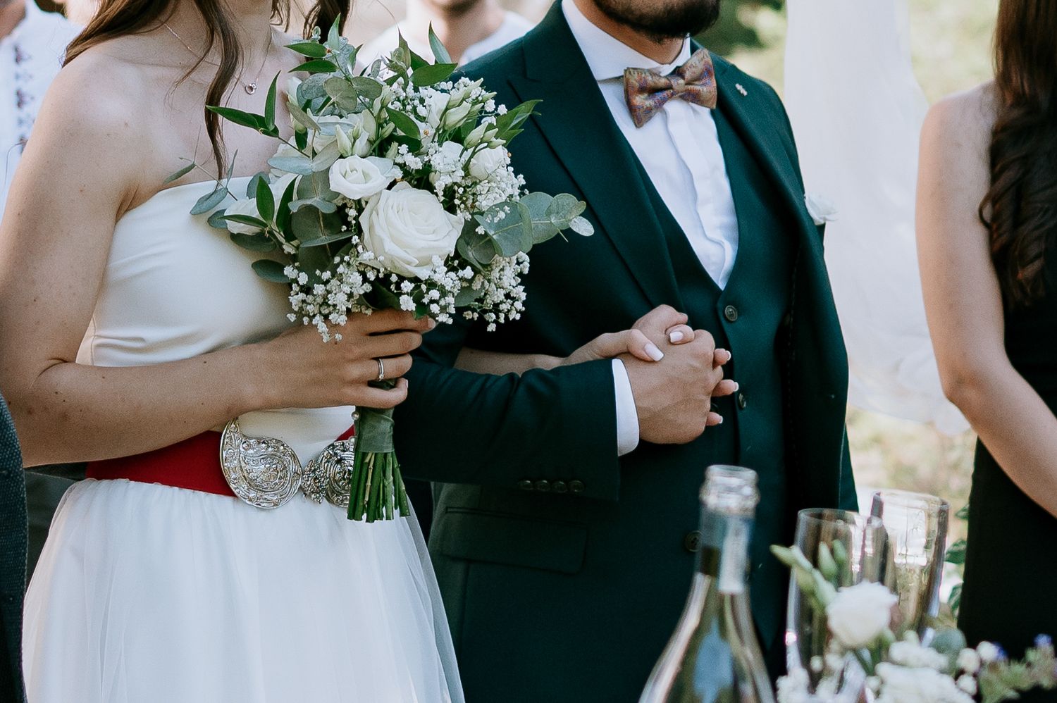 Wedding party gathers outdoors near white stone building with festive decorations and floral arrangements.