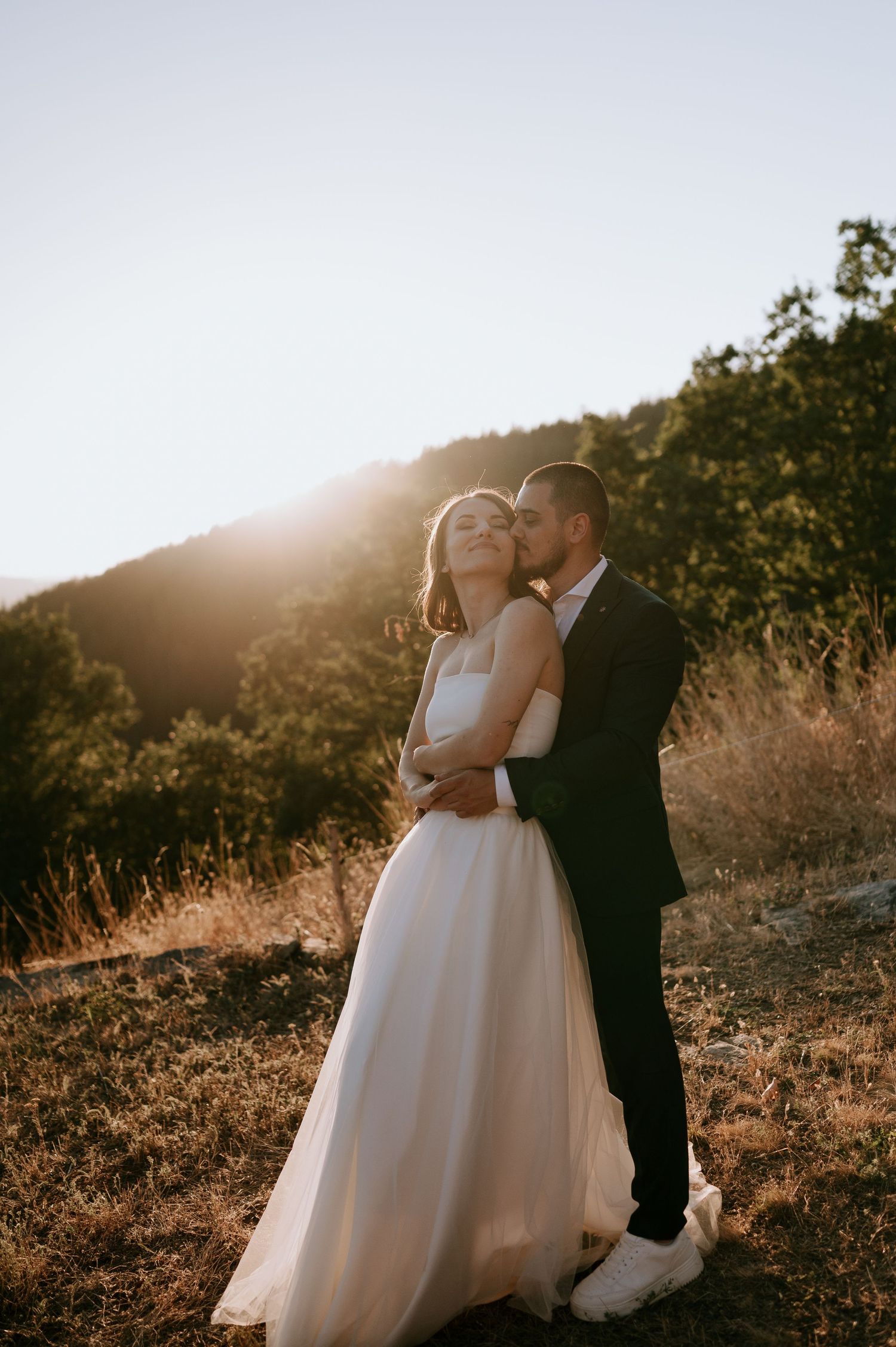 A romantic wedding couple embraces during golden hour on a hillside with sunlight streaming through the trees.