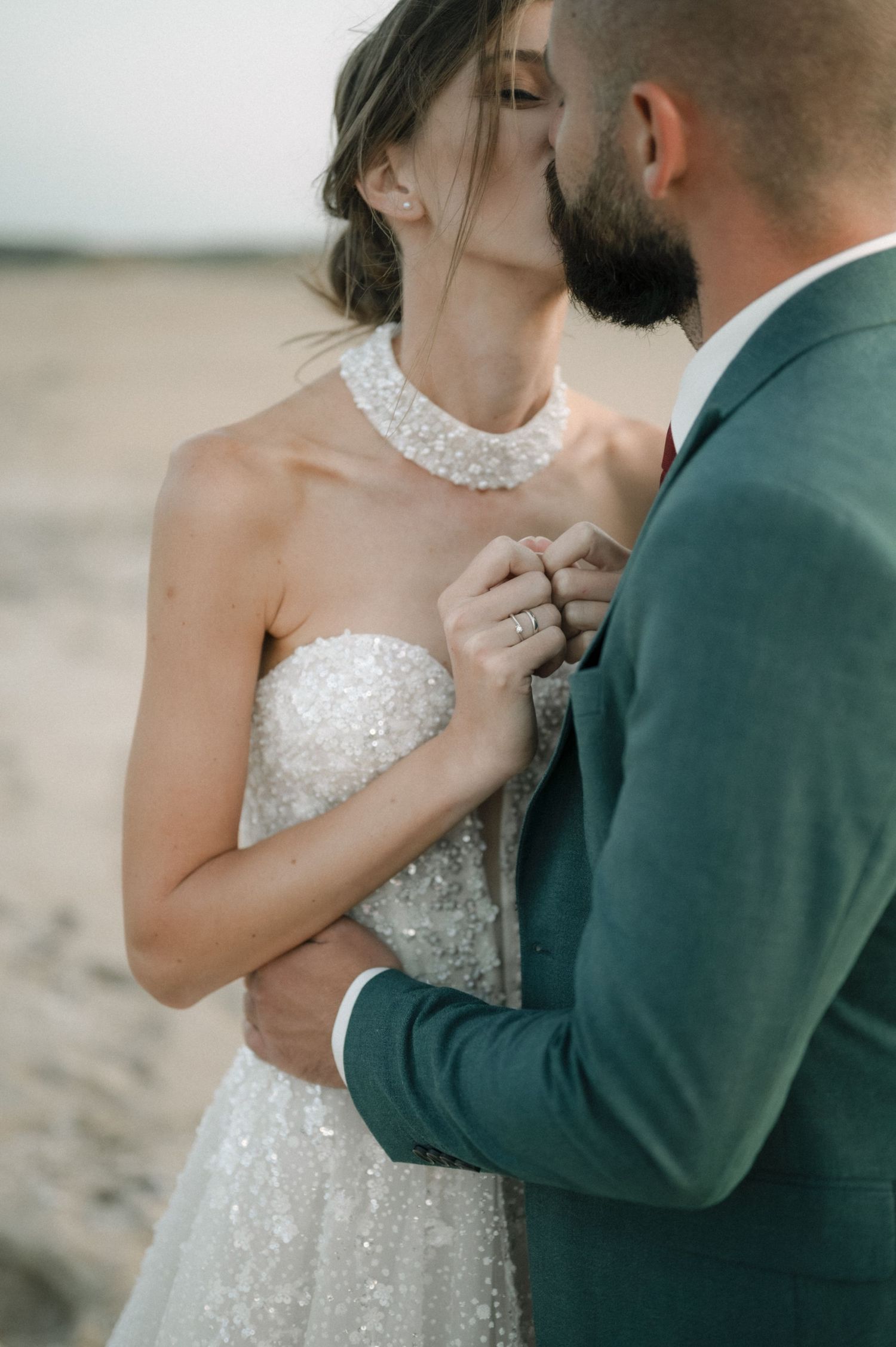 Bride in sequined strapless gown with pearl necklace shares tender moment with groom in teal suit on sandy beach.