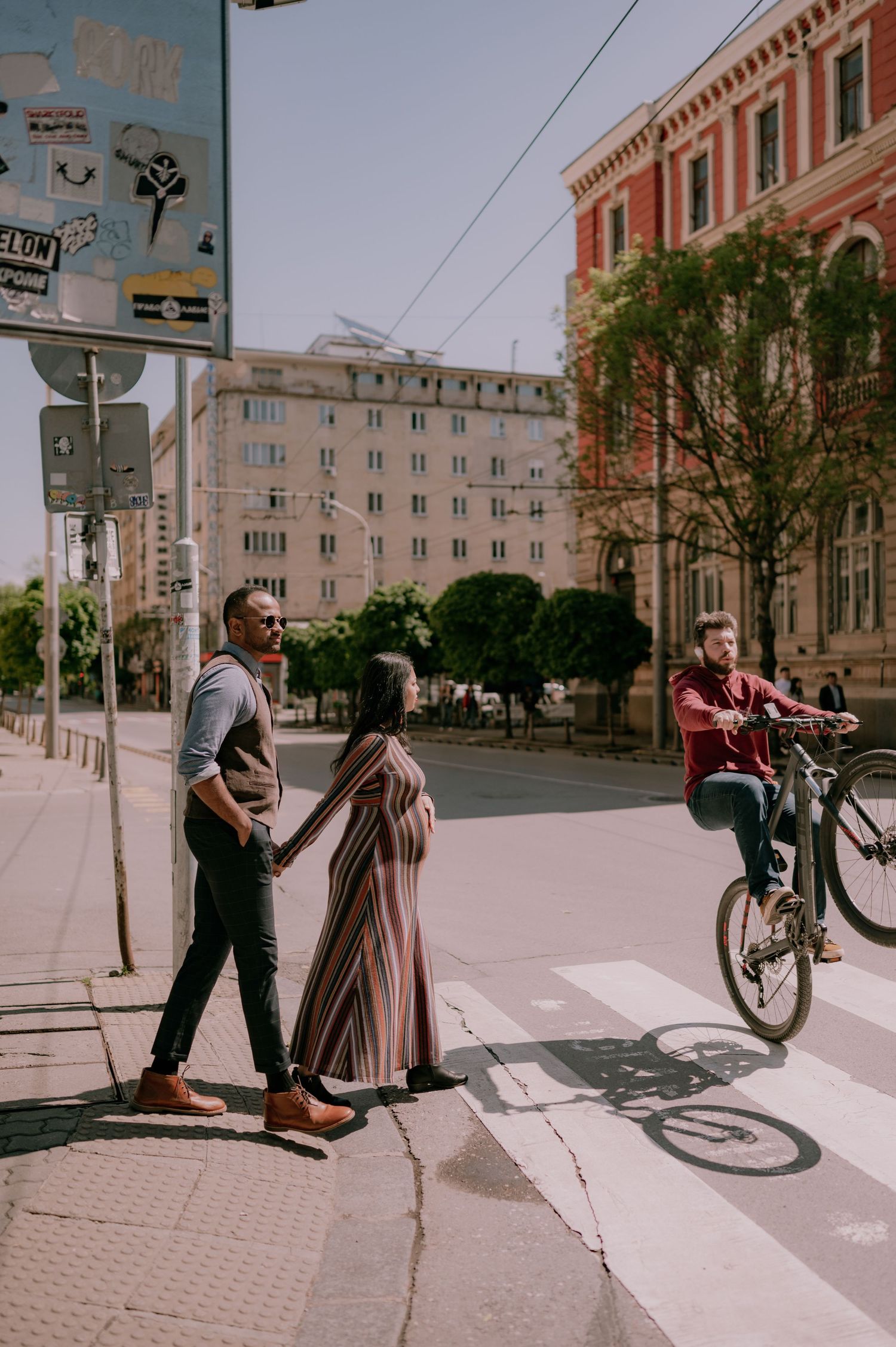 Couple holding hands at a city crosswalk on a sunny day, with a cyclist and urban buildings in the background.