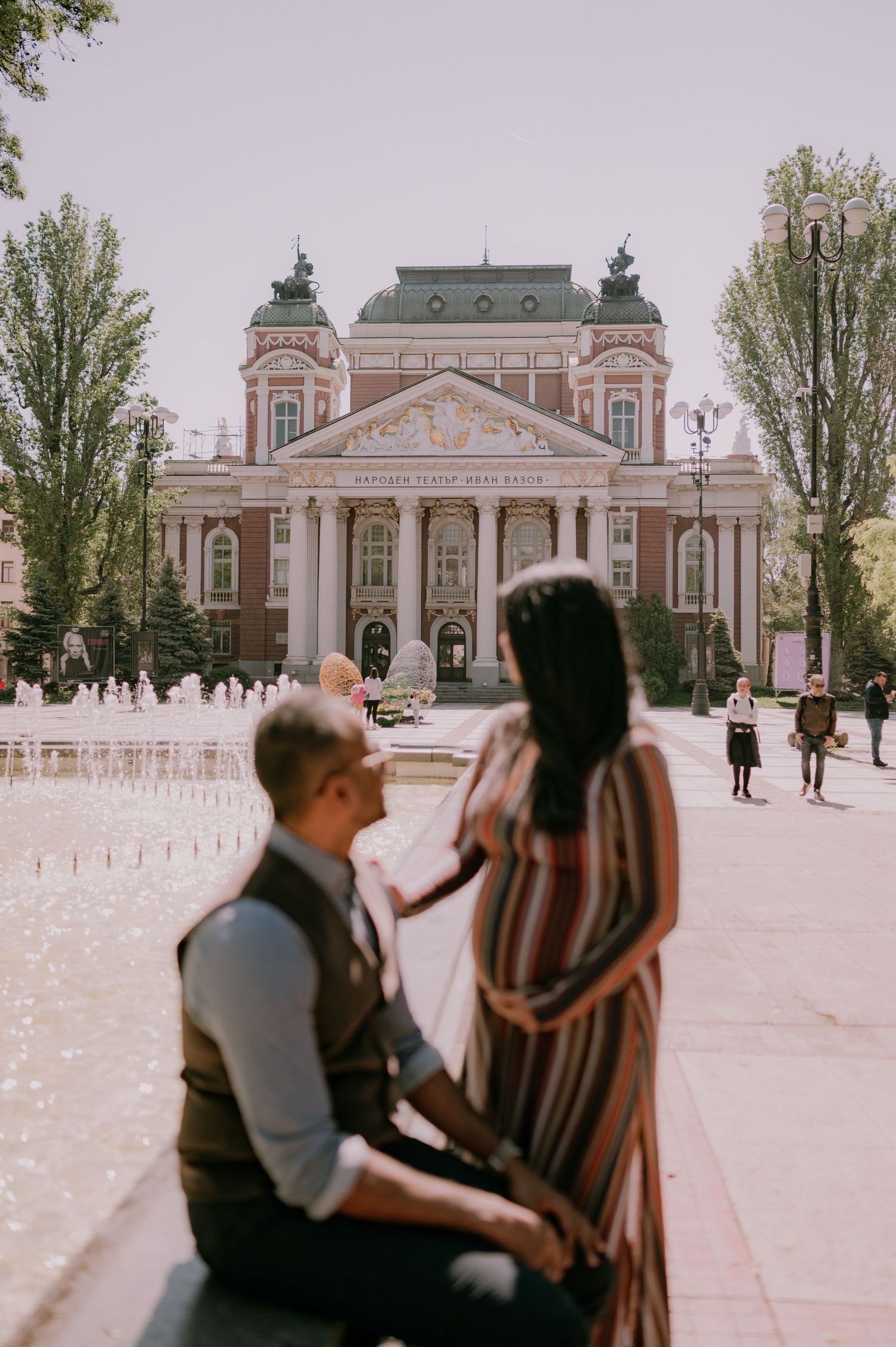 Couple in front of ornate pink historic building with fountain in Sofia, Bulgaria on a sunny day.