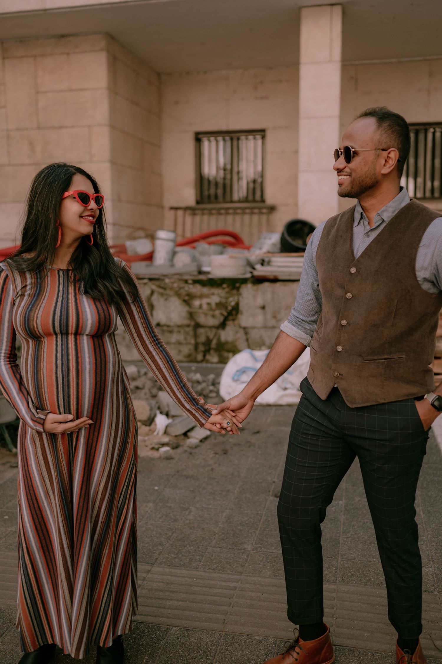 Pregnant woman in striped dress holds hands with smiling man in vest and sunglasses outdoors.