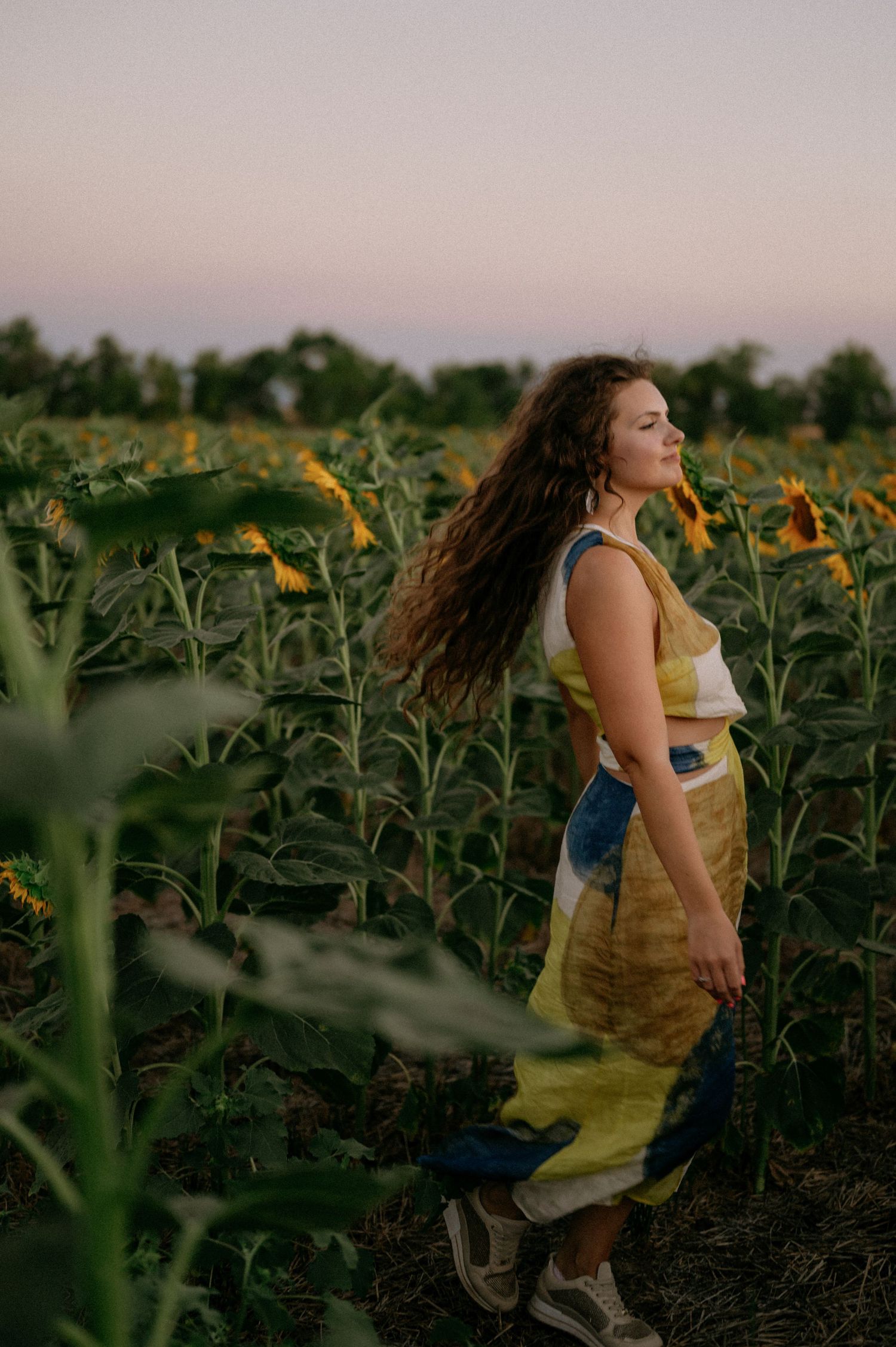 Woman in yellow tie-dye outfit standing in a sunflower field at dusk with long curly hair blowing in the breeze.