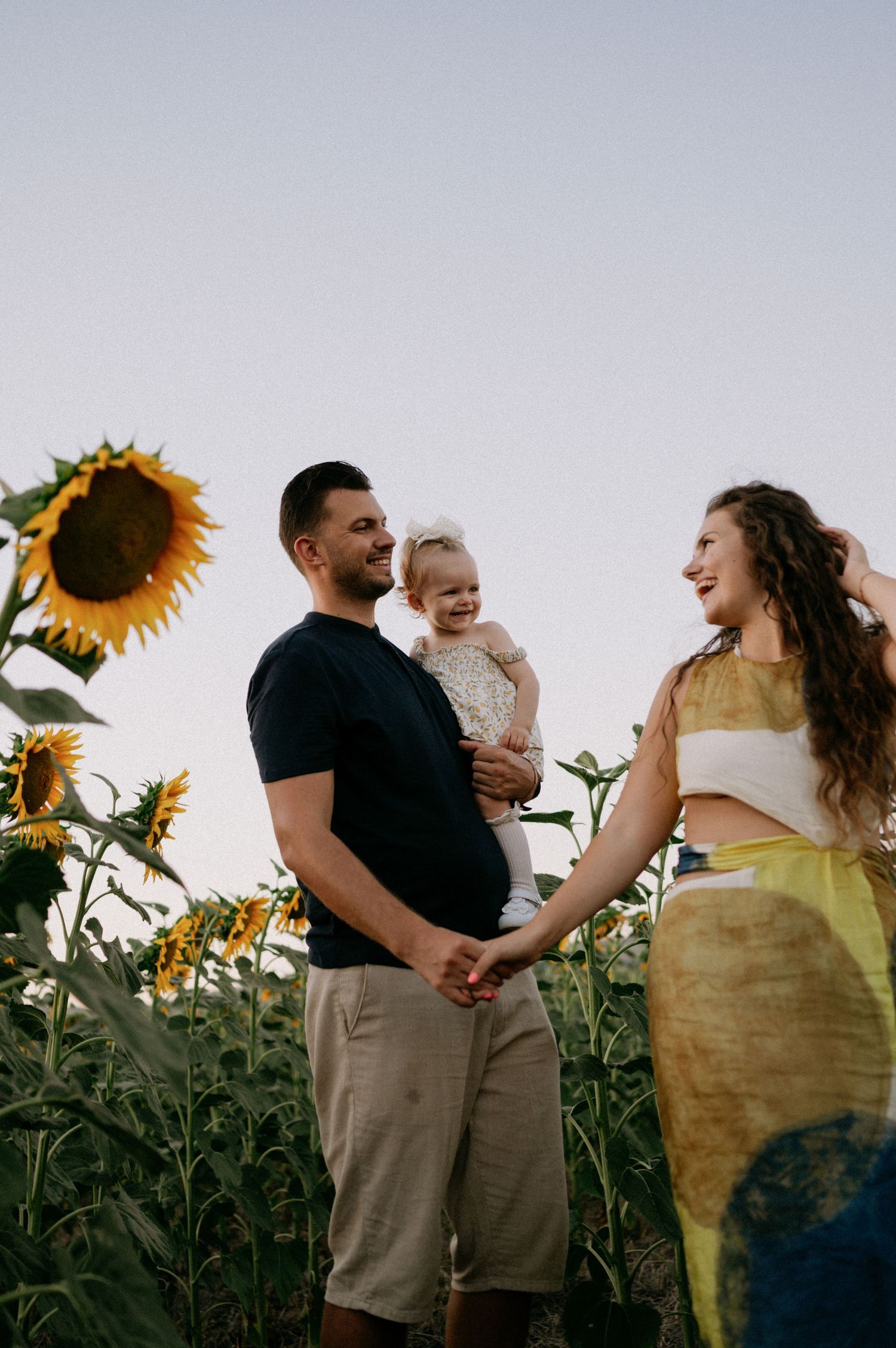 A happy family with a baby poses in a sunflower field at sunset, mom in yellow skirt, dad in black tee.