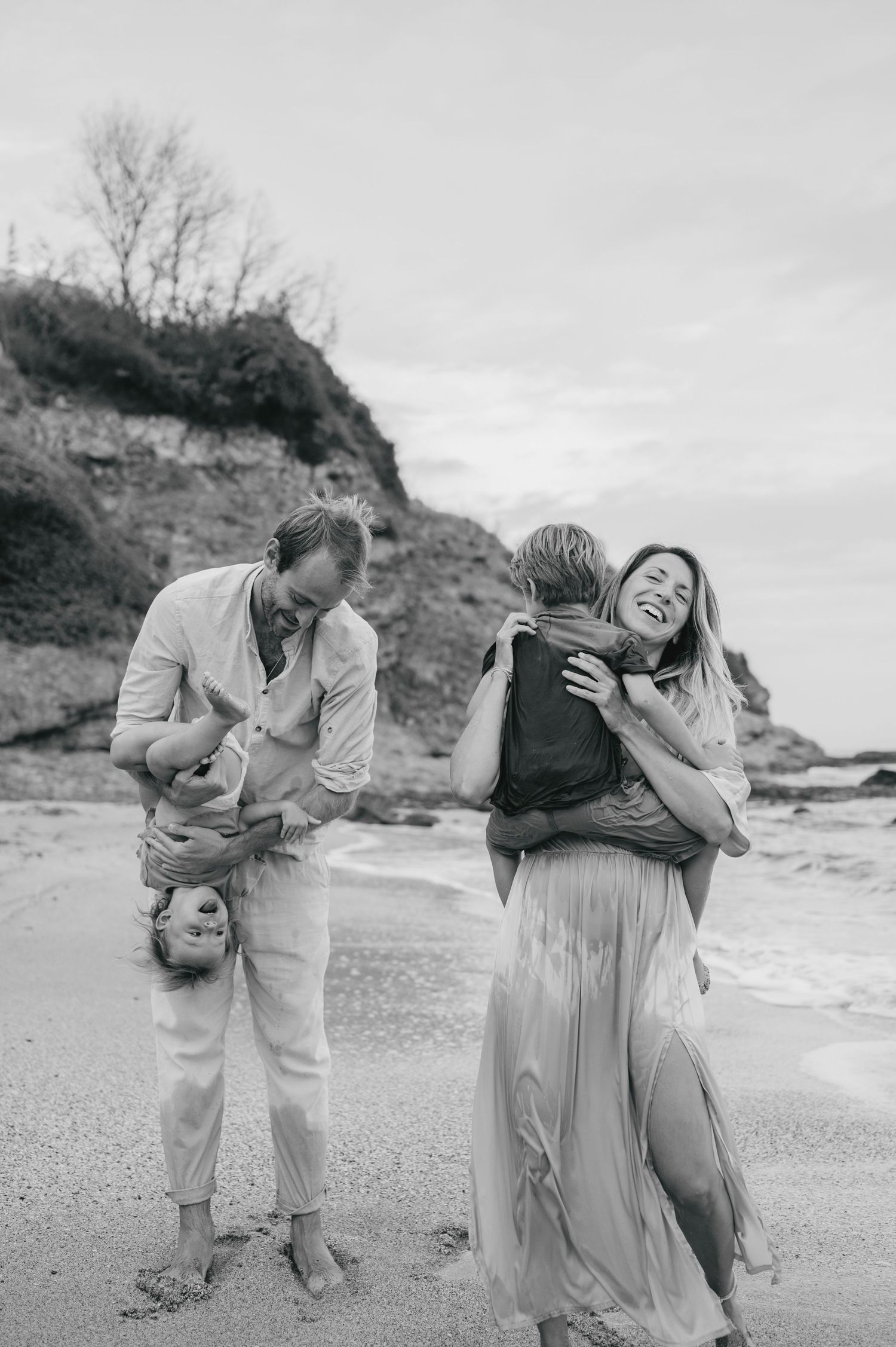Black and white beach family photo with parents playfully holding children, laughing on sandy shore with rocky cliffs behind.
