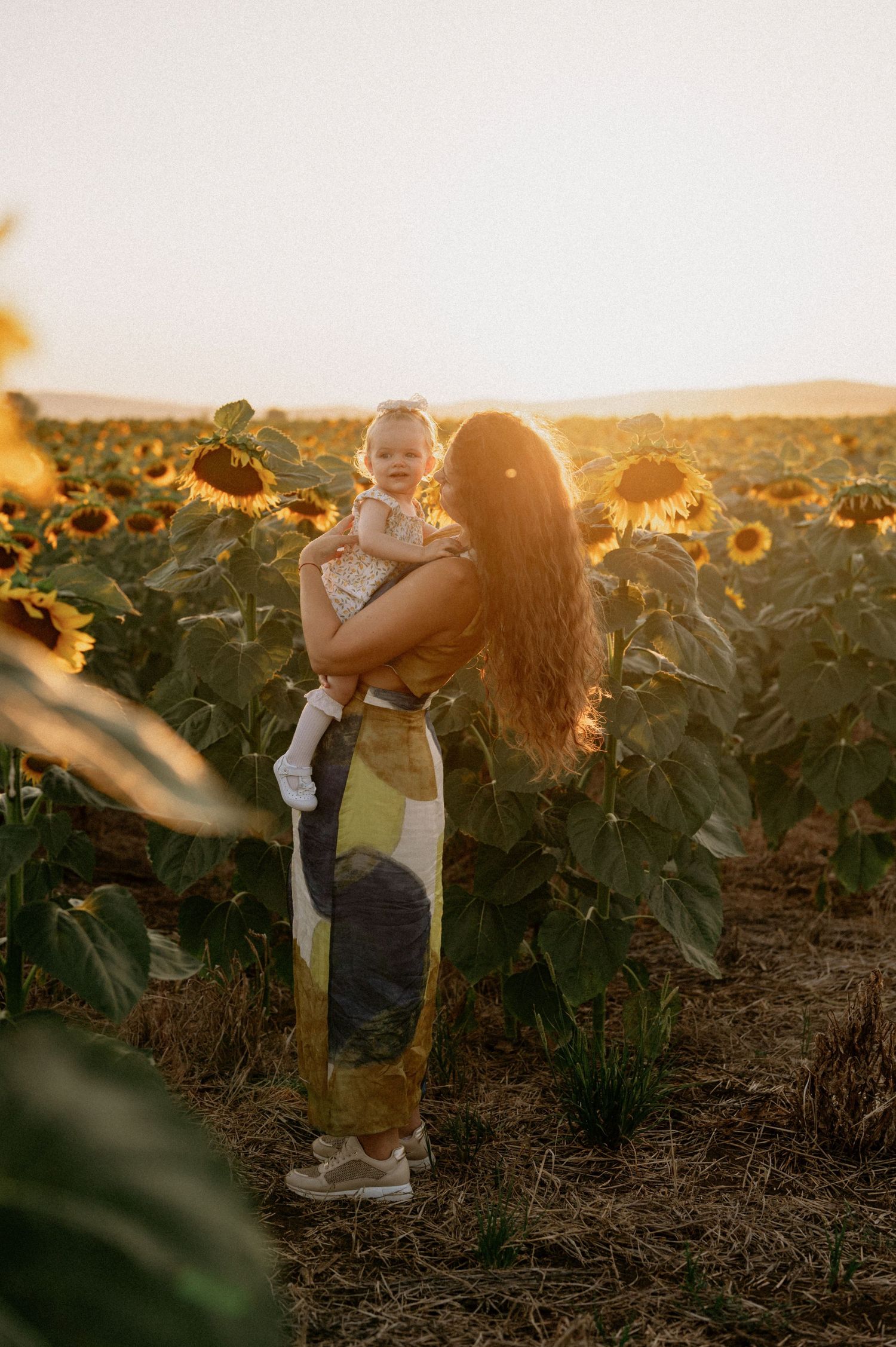 Mother holding baby in sunflower field at golden hour sunset, wearing floral dress with long hair.