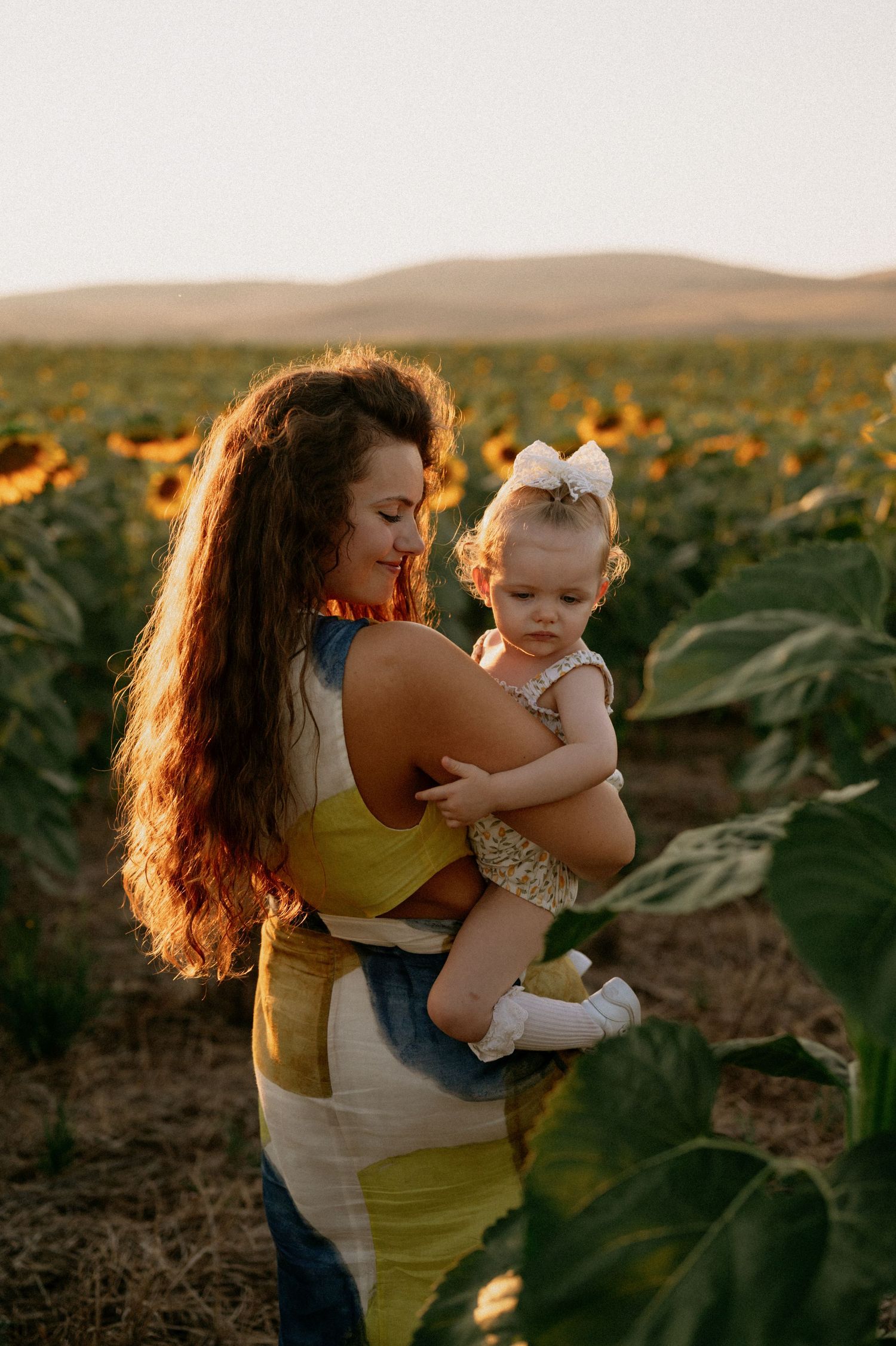 A mother holds her baby girl in a sunflower field at golden hour, both wearing summer outfits.