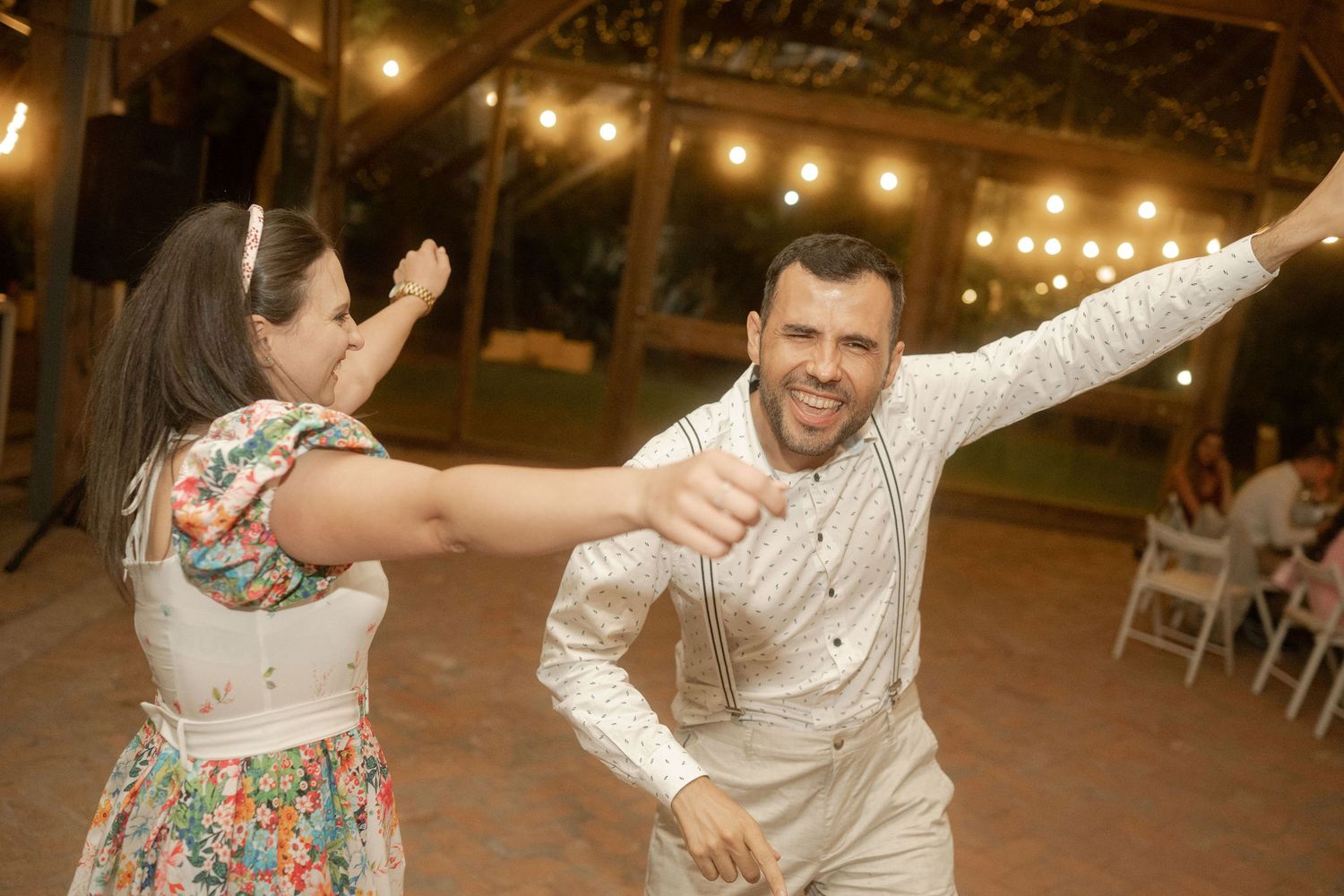 Two people dancing energetically at an outdoor evening reception with string lights overhead.