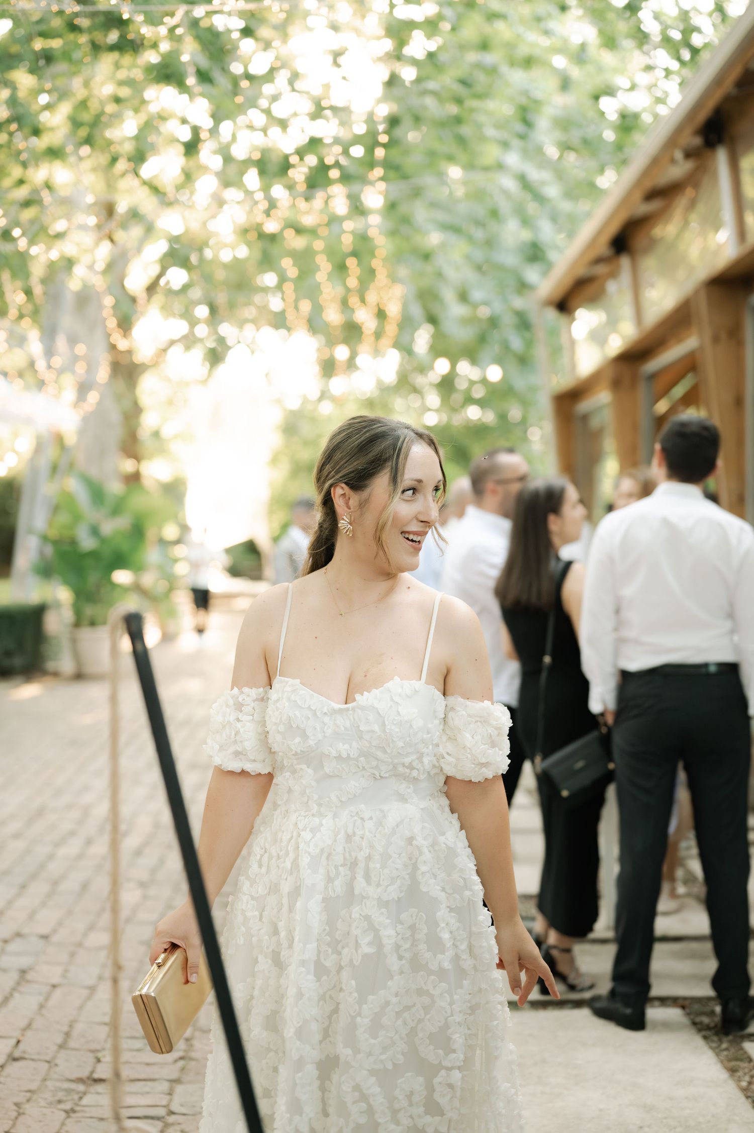 A bride in an off-shoulder lace wedding dress smiles while holding a clutch purse at an outdoor wedding venue.