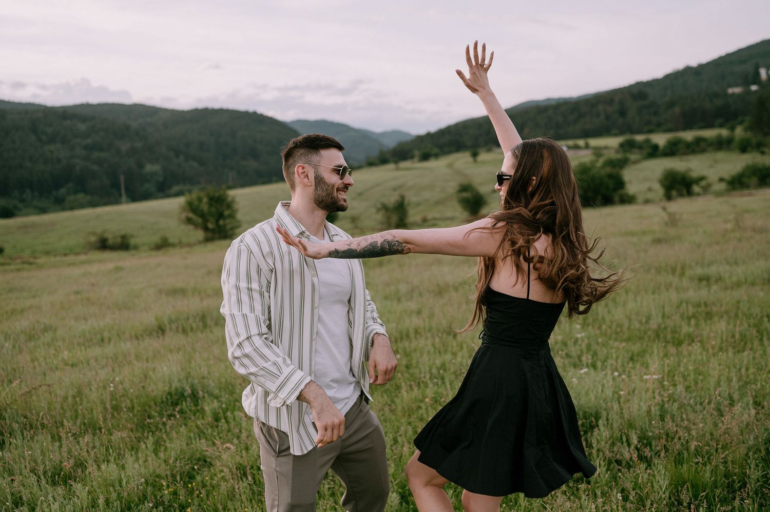 Joyful couple dances together in a black dress and striped shirt on a green hillside meadow under cloudy skies.