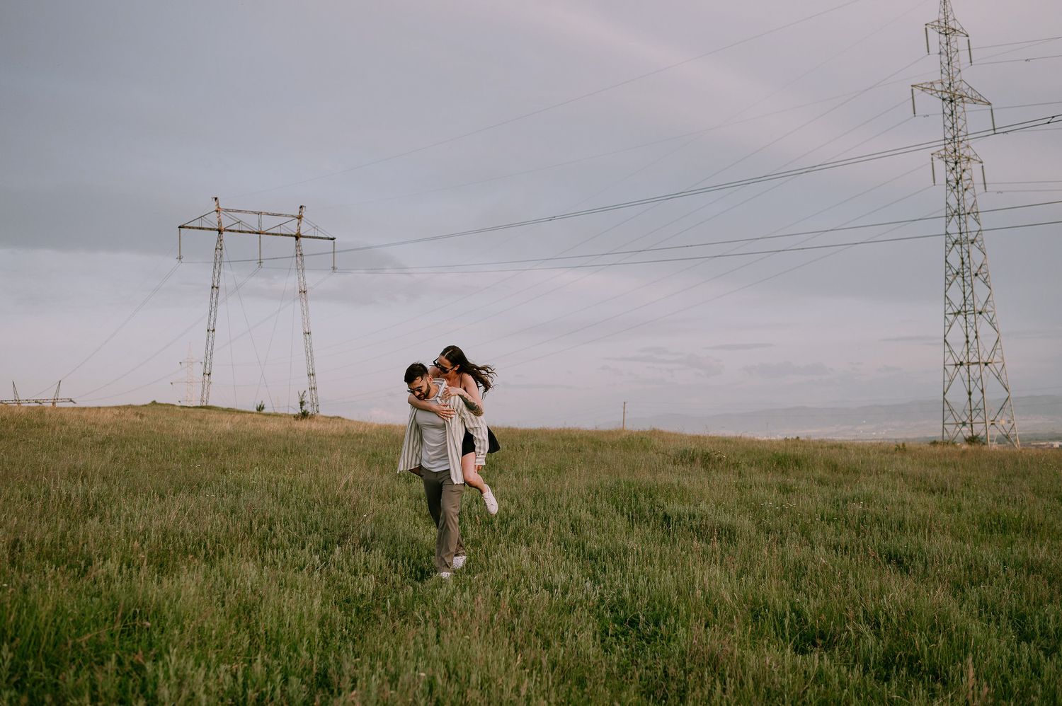 Two figures embrace while walking through a grassy field beneath power lines and transmission towers on an overcast day.