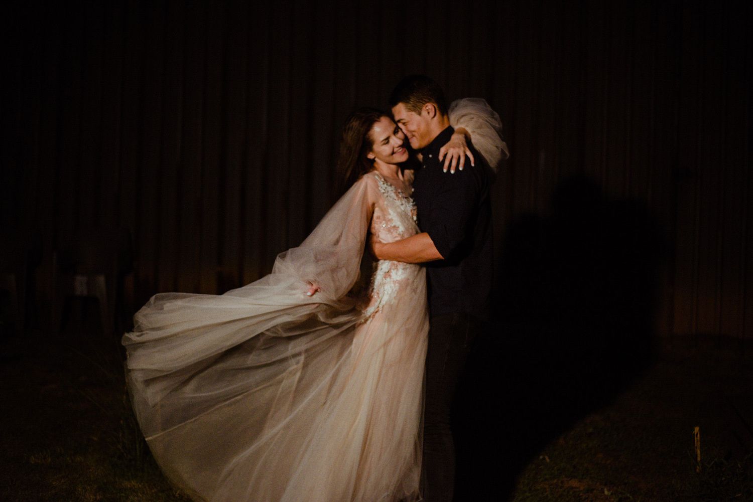 A romantic nighttime wedding portrait shows a couple embracing in flowing white dress and dark suit against a shadowy background.