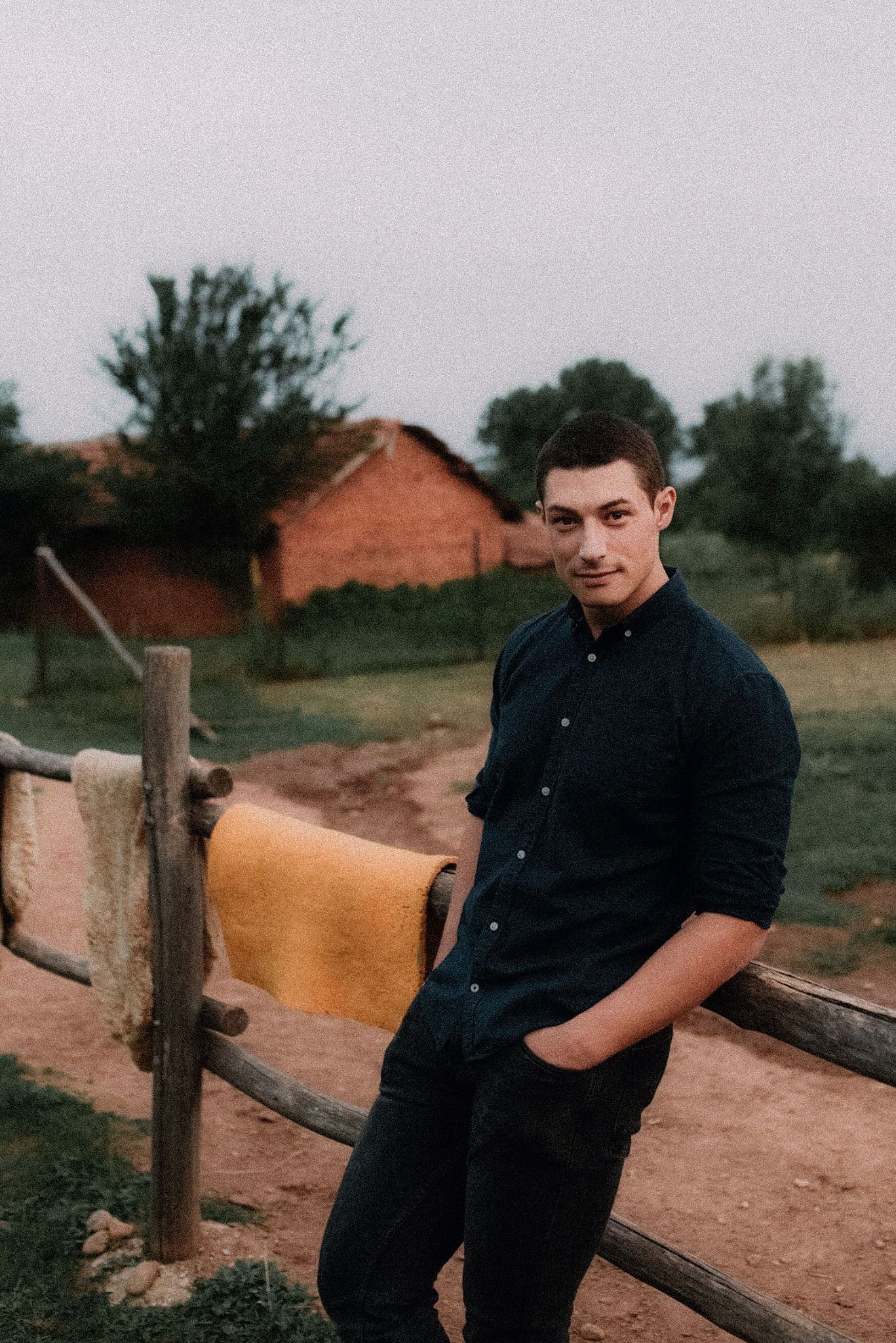 A person in dark clothing sits casually on a wooden fence in a rural setting with reddish buildings visible in background.