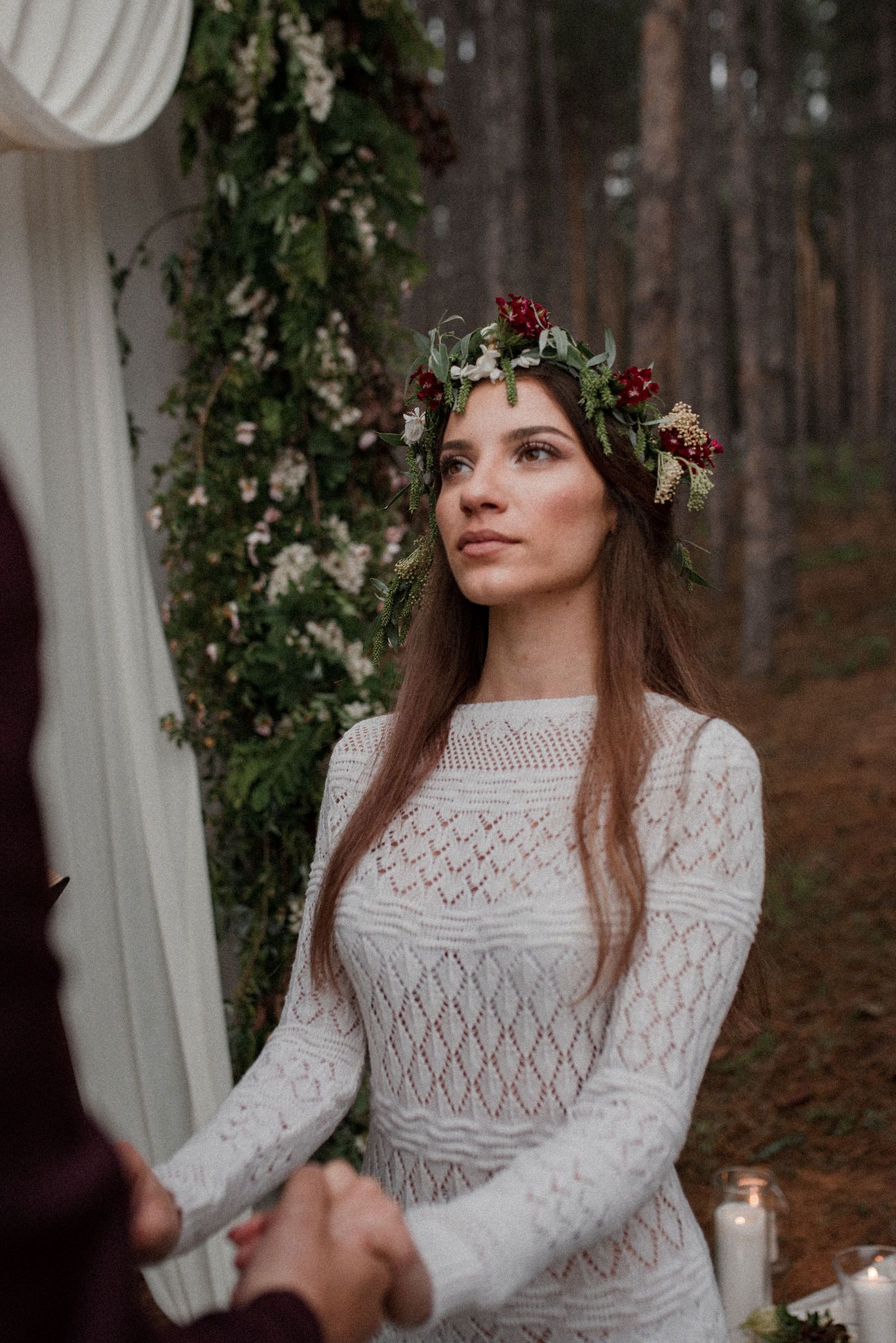 A person wearing a white lace dress and floral crown poses in front of a forest backdrop in a romantic outdoor setting.