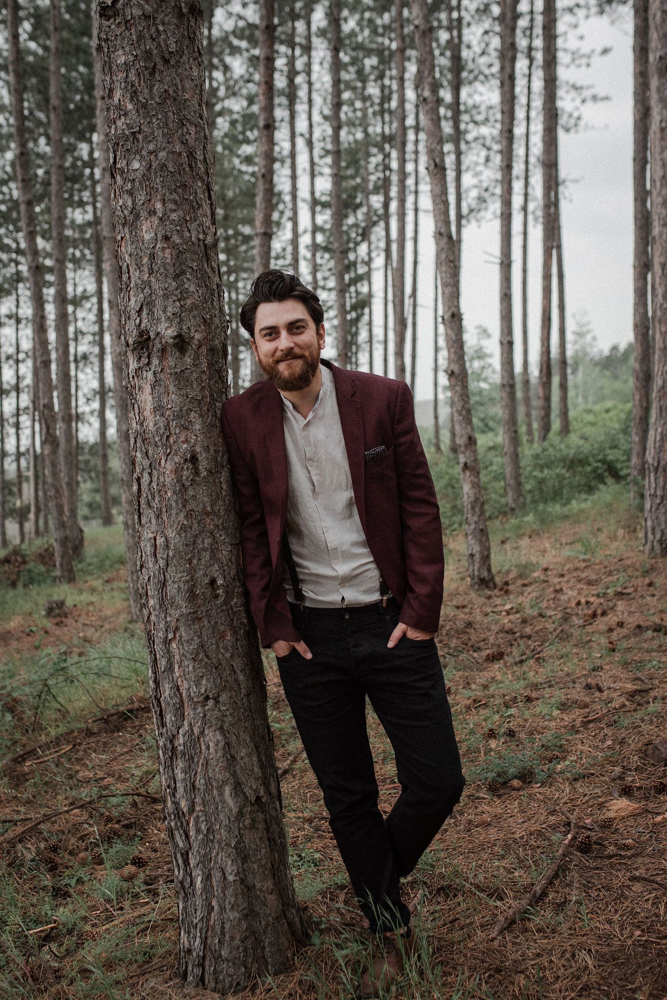 A person in a burgundy blazer and white shirt poses casually against pine trees in a forest setting.