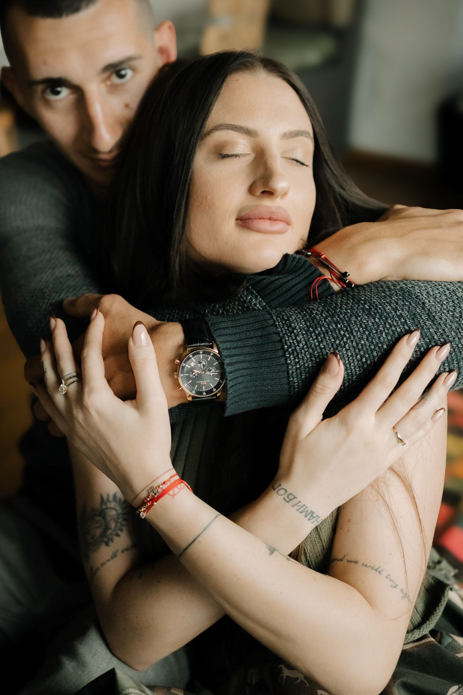 A couple embraces in a tender moment wearing dark sweaters and jewelry in a dimly lit setting.