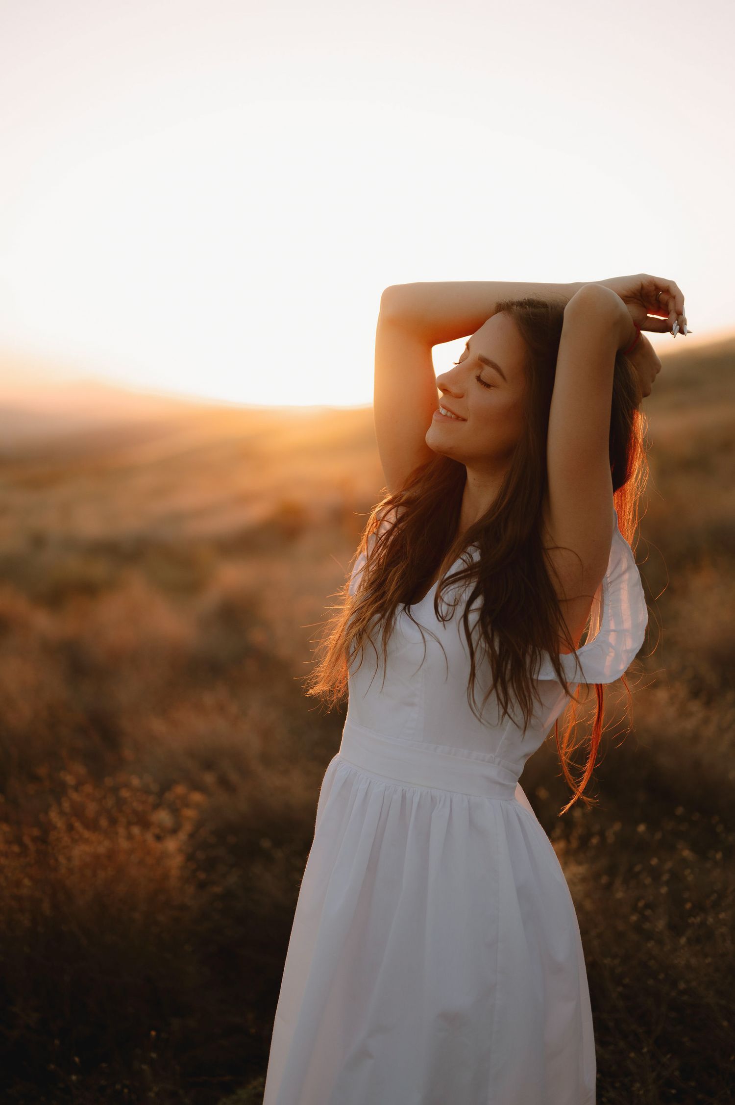 A figure in a flowing white dress poses gracefully against a golden sunset in a rustic field.