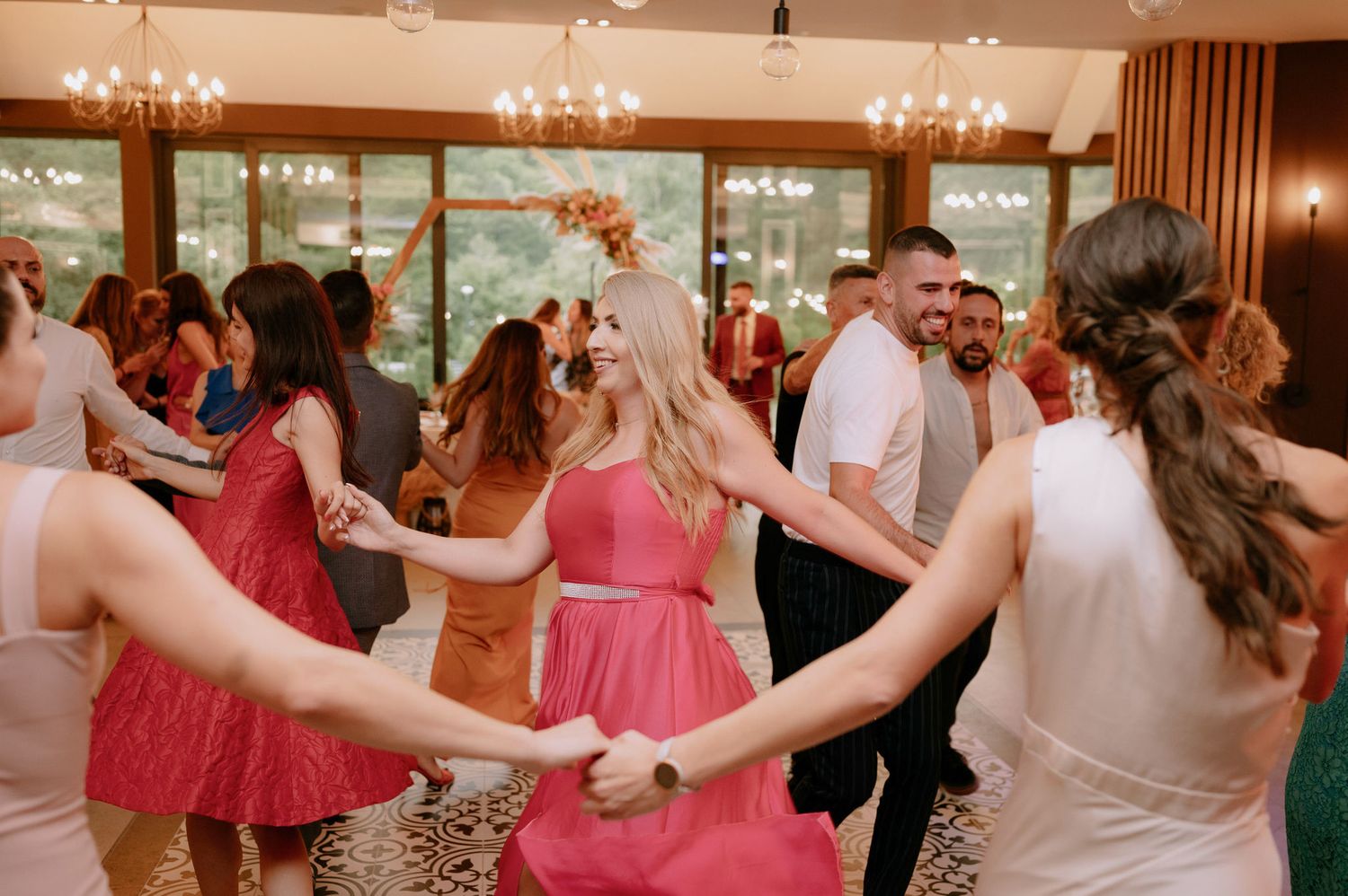 Wedding guests dance and celebrate on a wooden dance floor at an indoor reception venue with warm lighting.