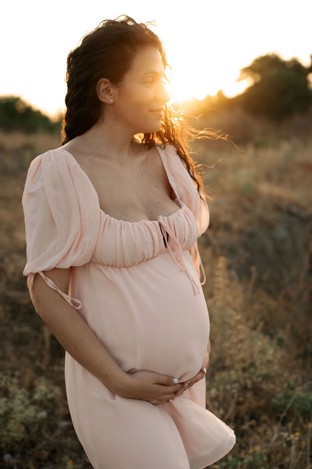 A woman in a flowy white dress cradles her pregnant belly during a sunset maternity photoshoot.