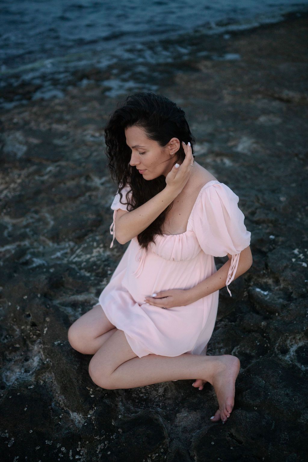 A figure in a soft pink dress sits contemplatively on dark rocks in a moody artistic photoshoot.