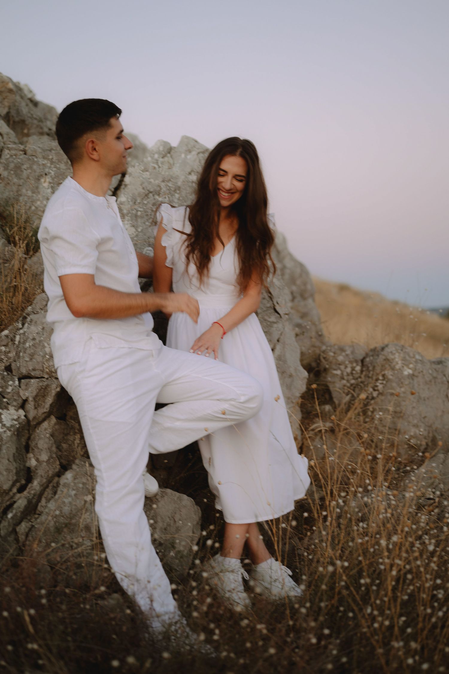 A couple in white outfits dances playfully among rocky terrain at sunset.
