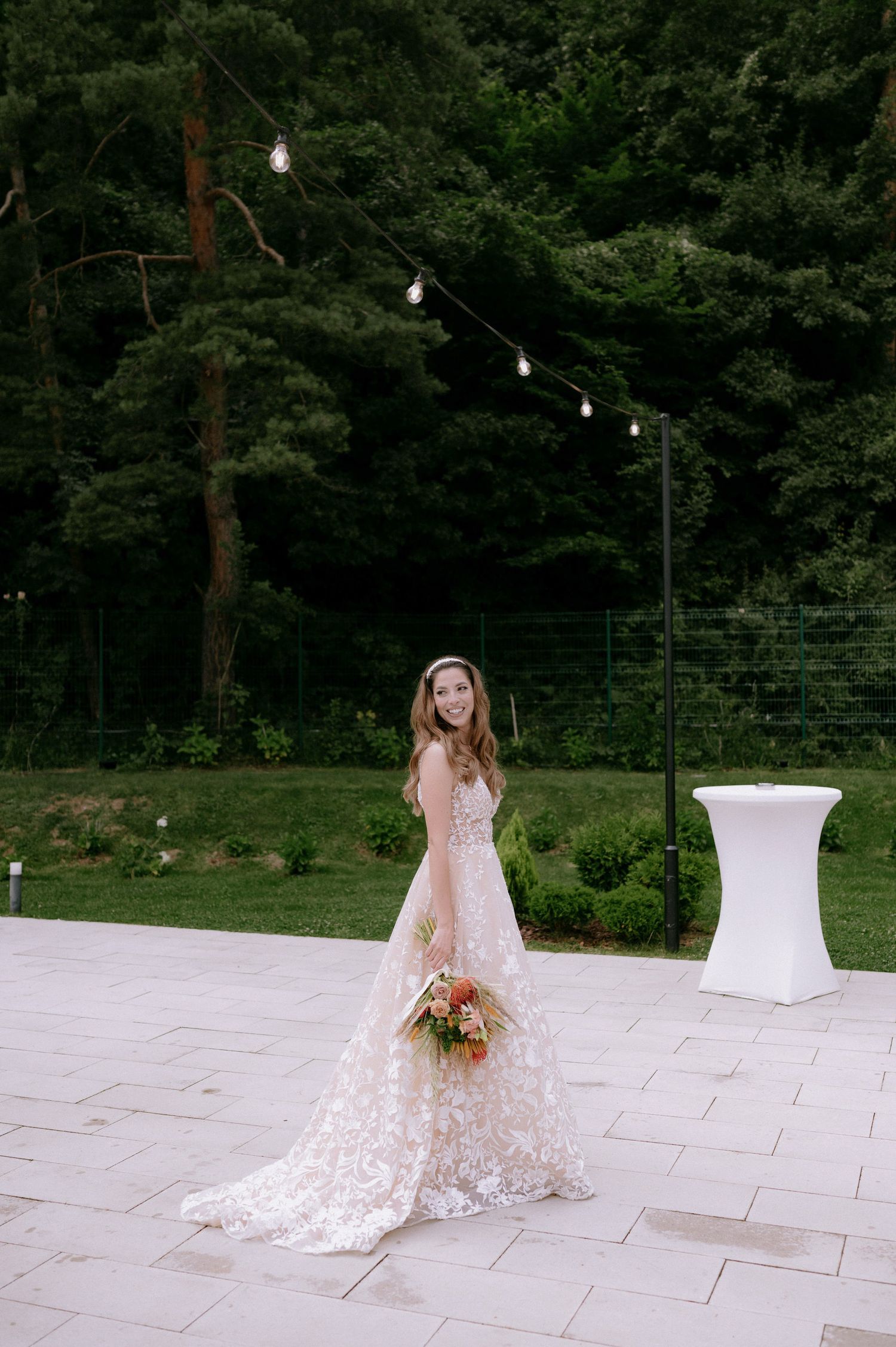 A bride in a delicate lace wedding gown poses gracefully on a white stone patio surrounded by lush greenery.
