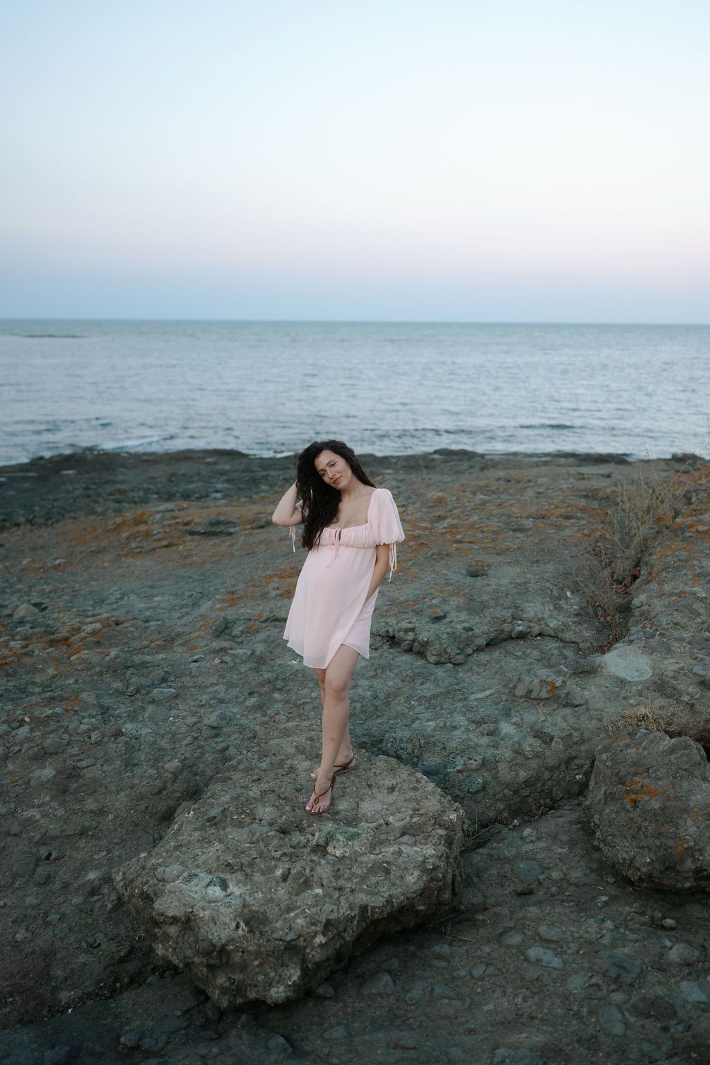 A person in a light dress stands on rocky coastal cliffs overlooking the ocean at dusk.