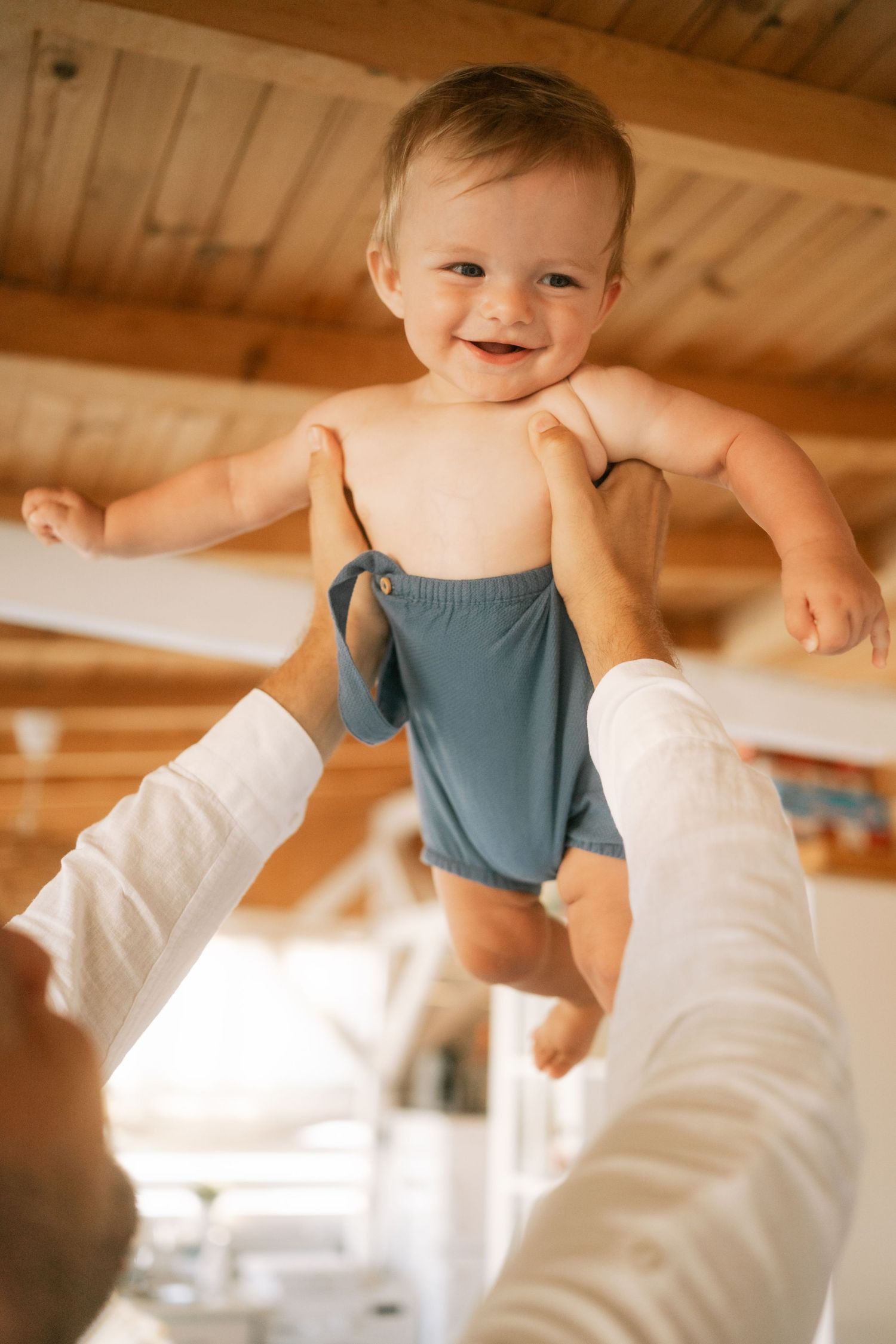 A series of photos showing a small baby in a blue romper being lifted playfully in the air against a wooden ceiling.