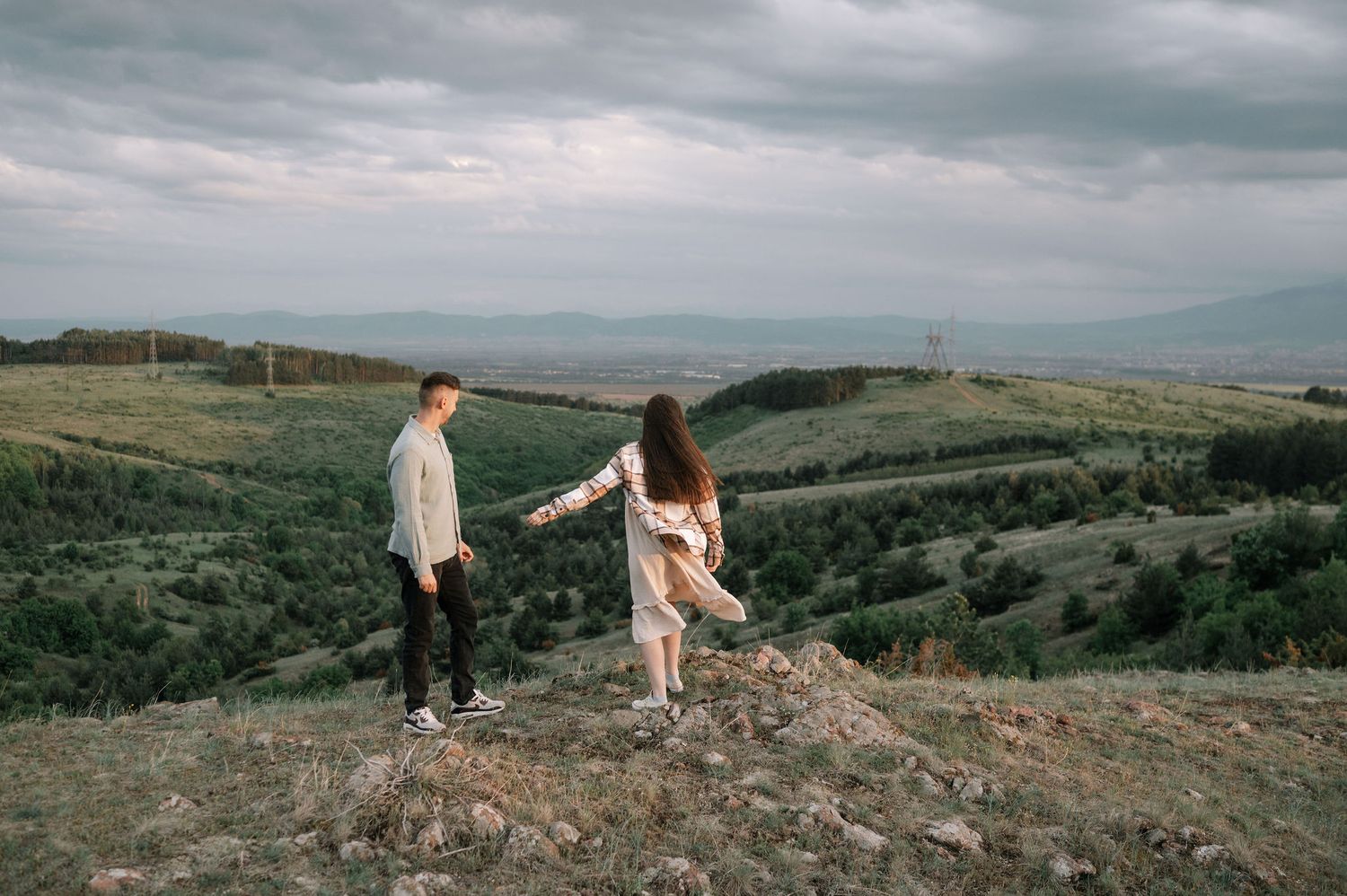 Two people walk along a scenic mountain ridge overlooking rolling green hills under a moody cloudy sky.