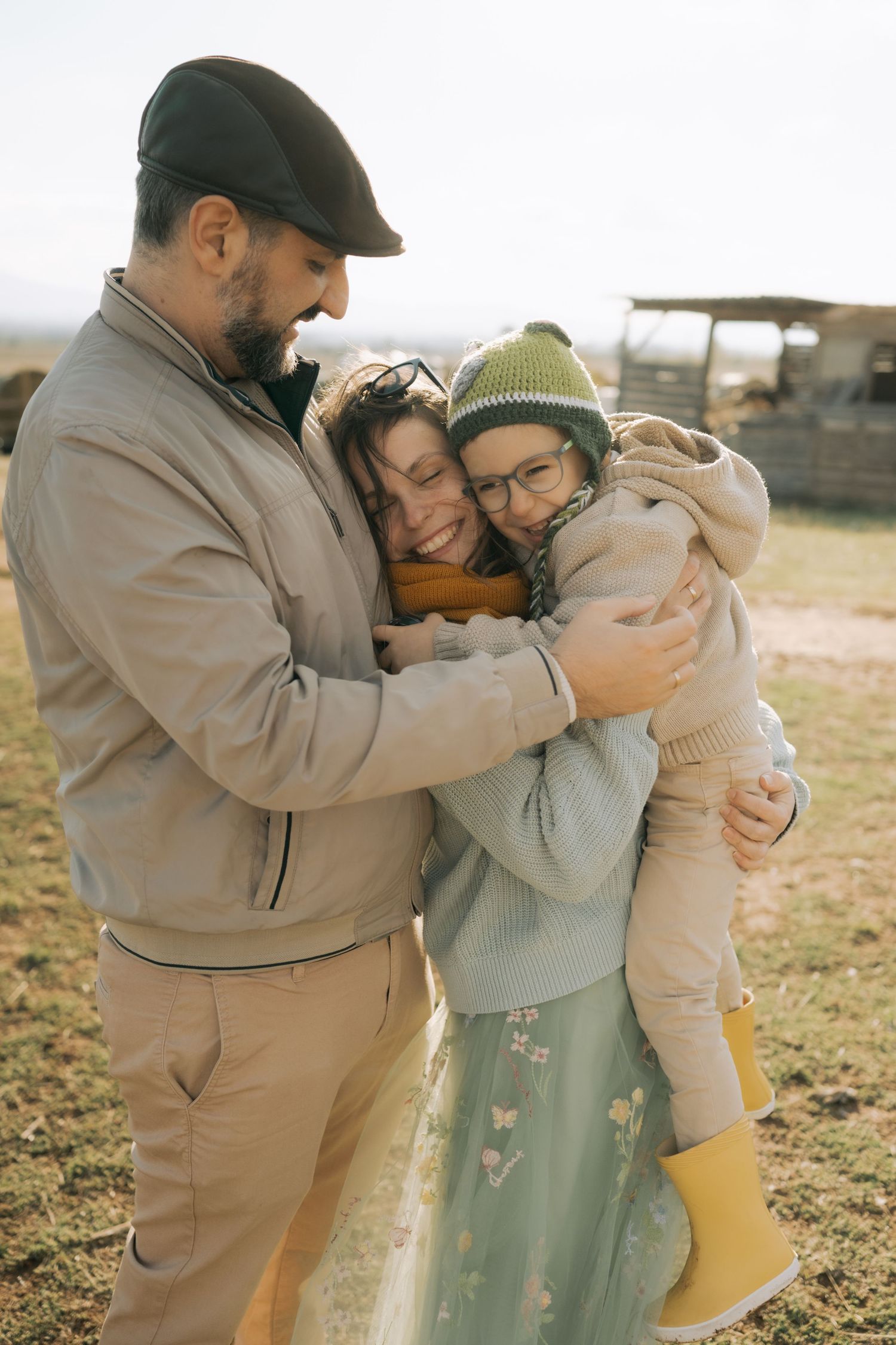 Family of three embraces outdoors in warm sunlight wearing casual clothing on grassy field.