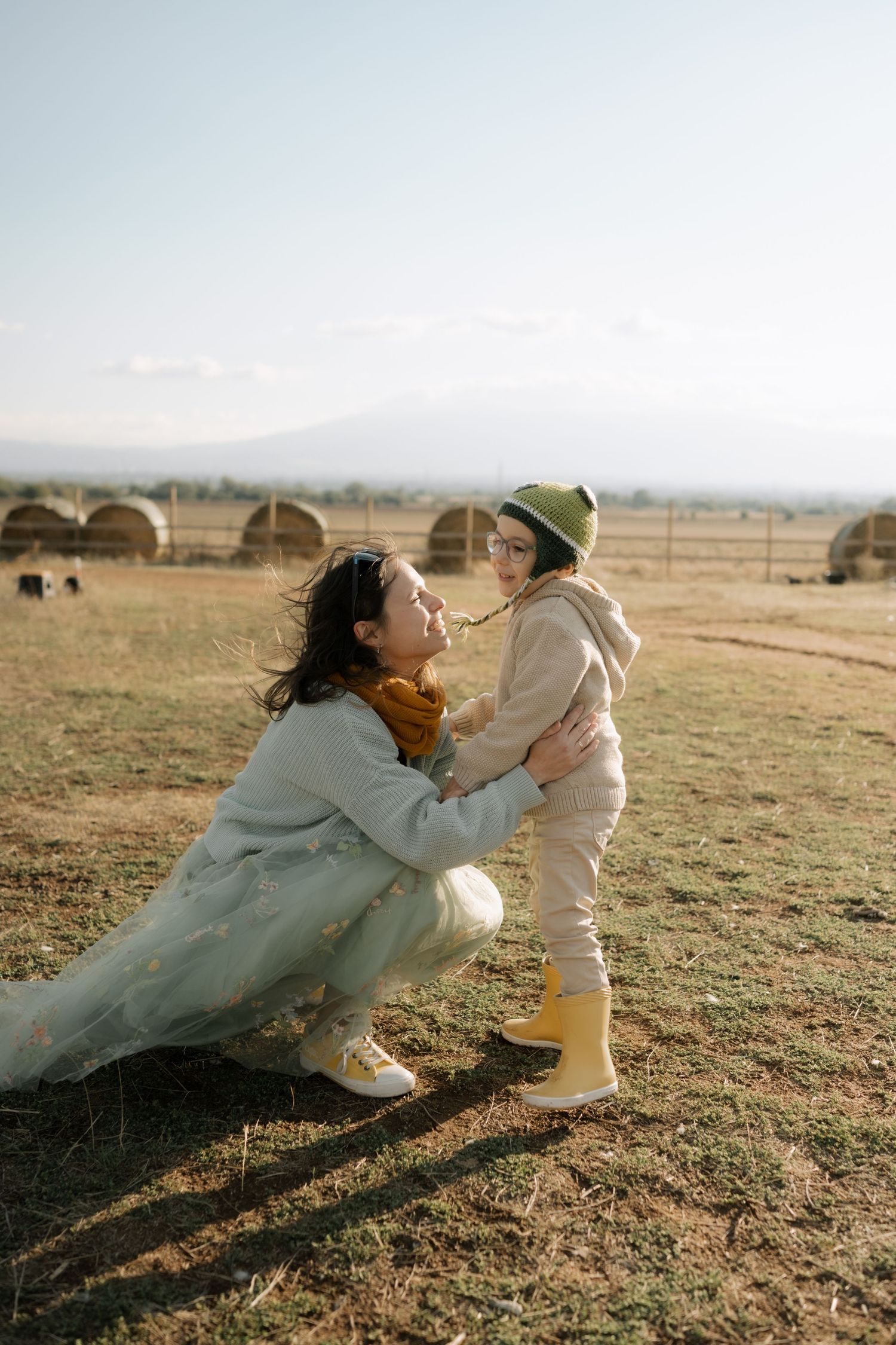 A mother in a flowing dress plays with a toddler in white on a grassy field at sunset, creating a tender moment.